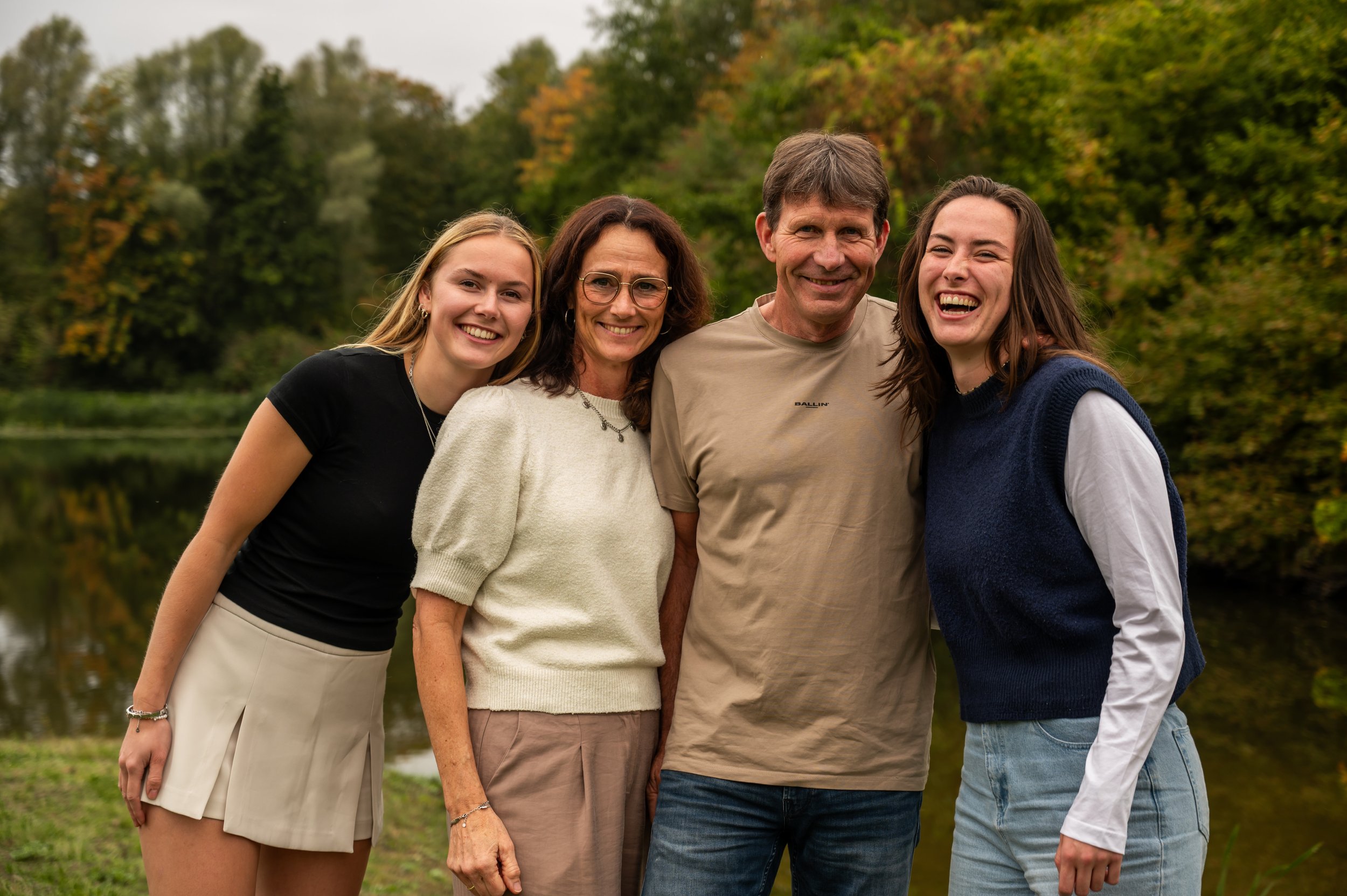 Vier mensen staan dicht bij elkaar buiten, lachend, met een bos en water achtergrond, in de herfst.