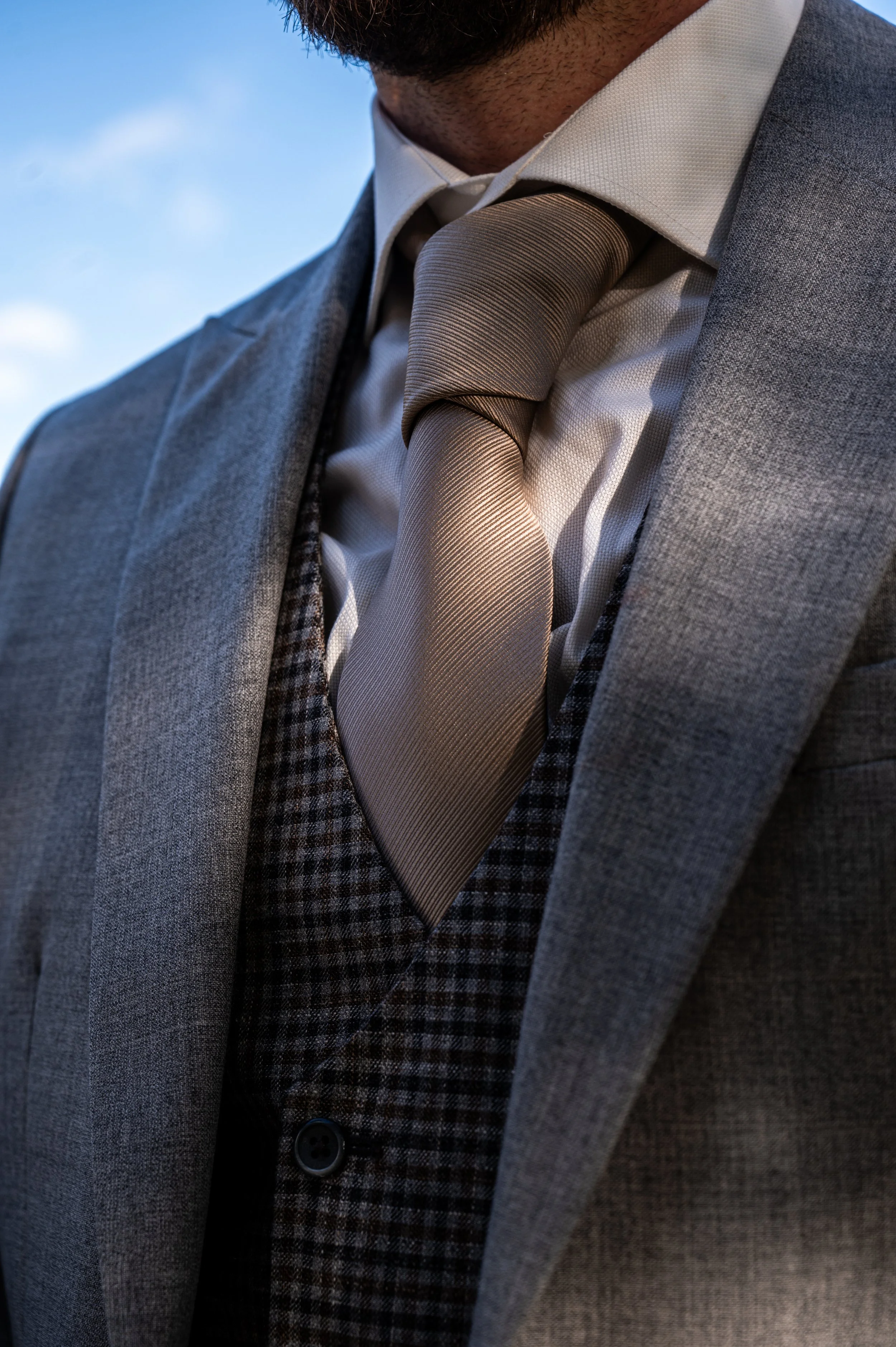 Close-up van een man in een grijs pak en beige stropdas, met een witte overhemd, tegen een blauwe lucht.