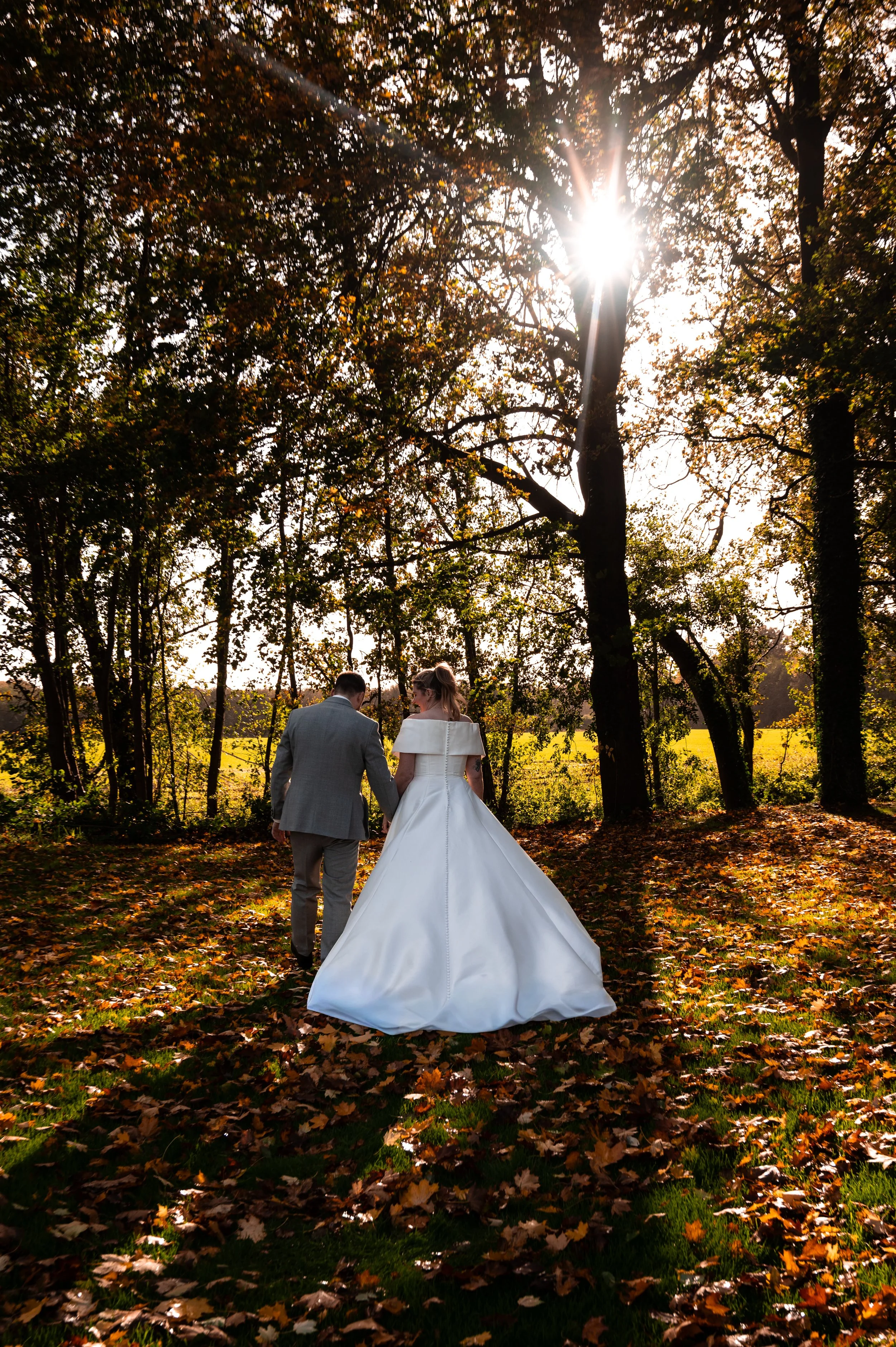 Een getrouwde paar wandelend door een bos in de herfst, met de zon die door de bomen schijnt, een vrouw in een witte trouwjurk en een man in een grijs pak.