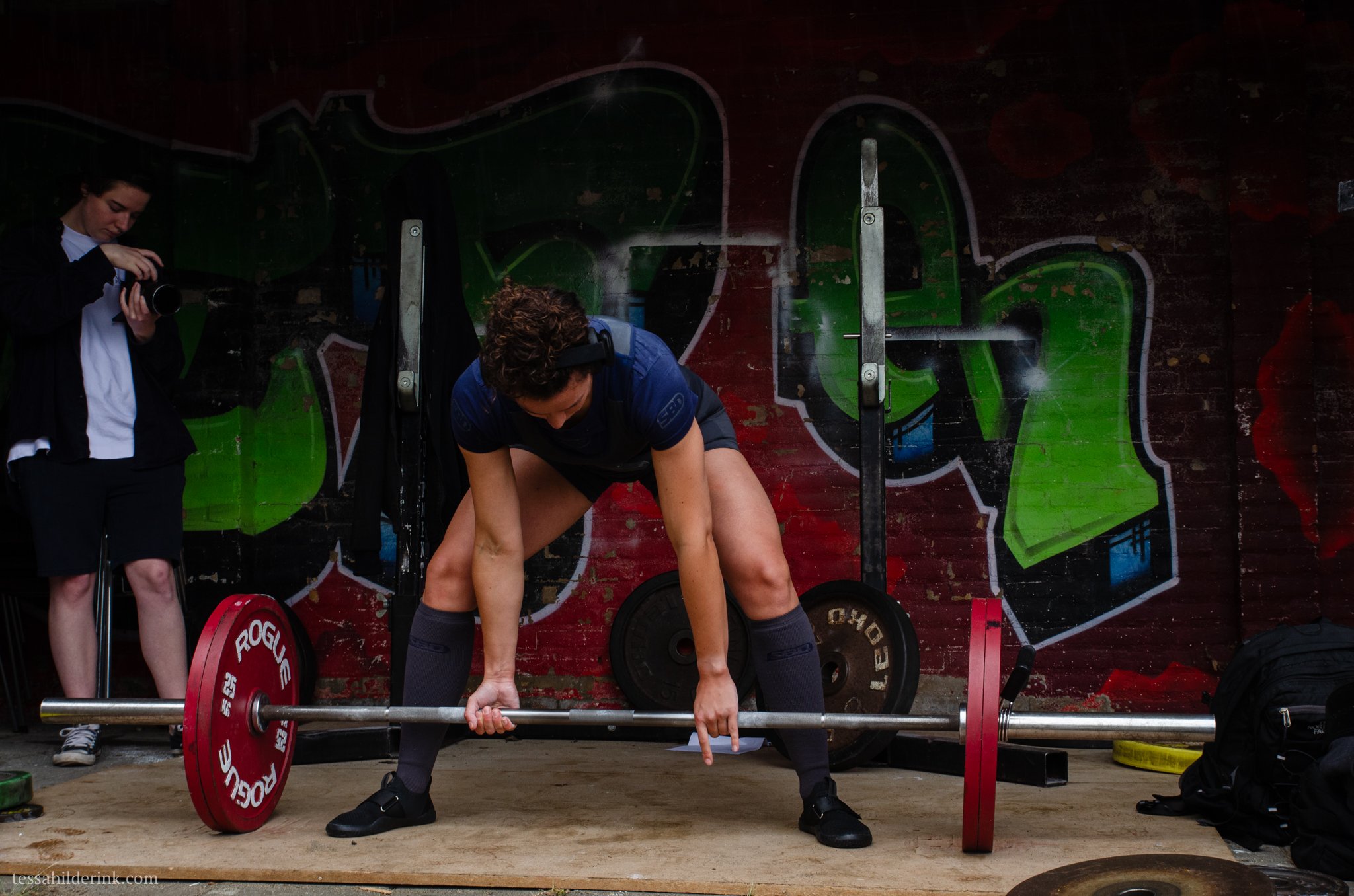 Person preparing to lift a barbell with red weight plates in a gym with graffiti on the wall and another person filming.