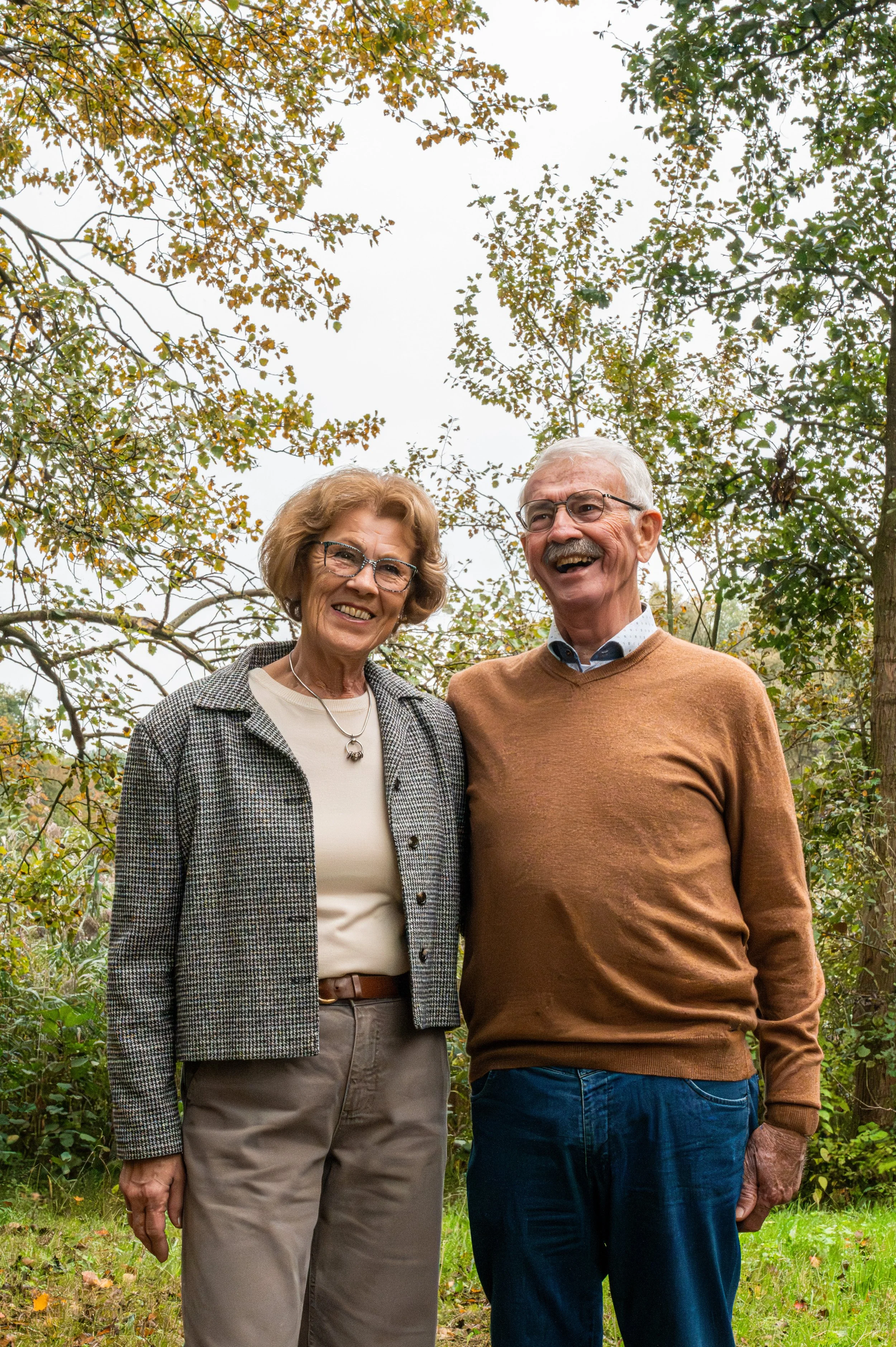 Een oudere vrouw en een oudere man staan samen buiten in een bos, lachend en poseren voor de camera.