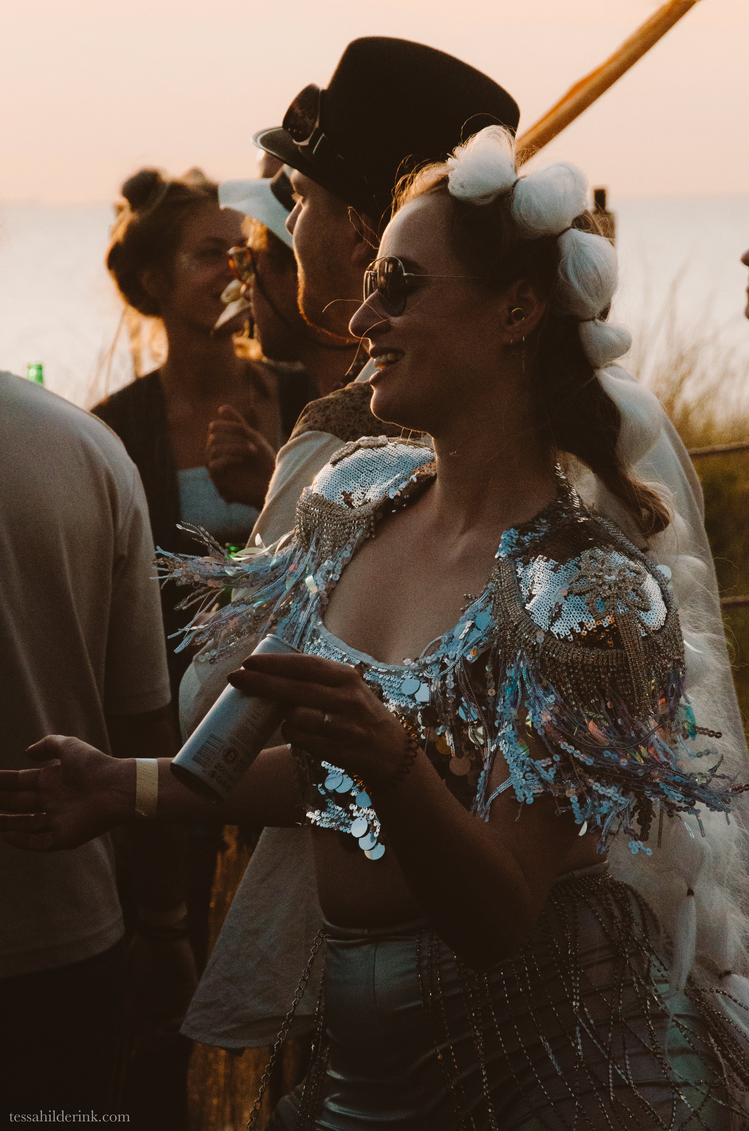 People at an outdoor event during sunset, with a woman wearing a sequined outfit and holding a can, and a man in a hat.
