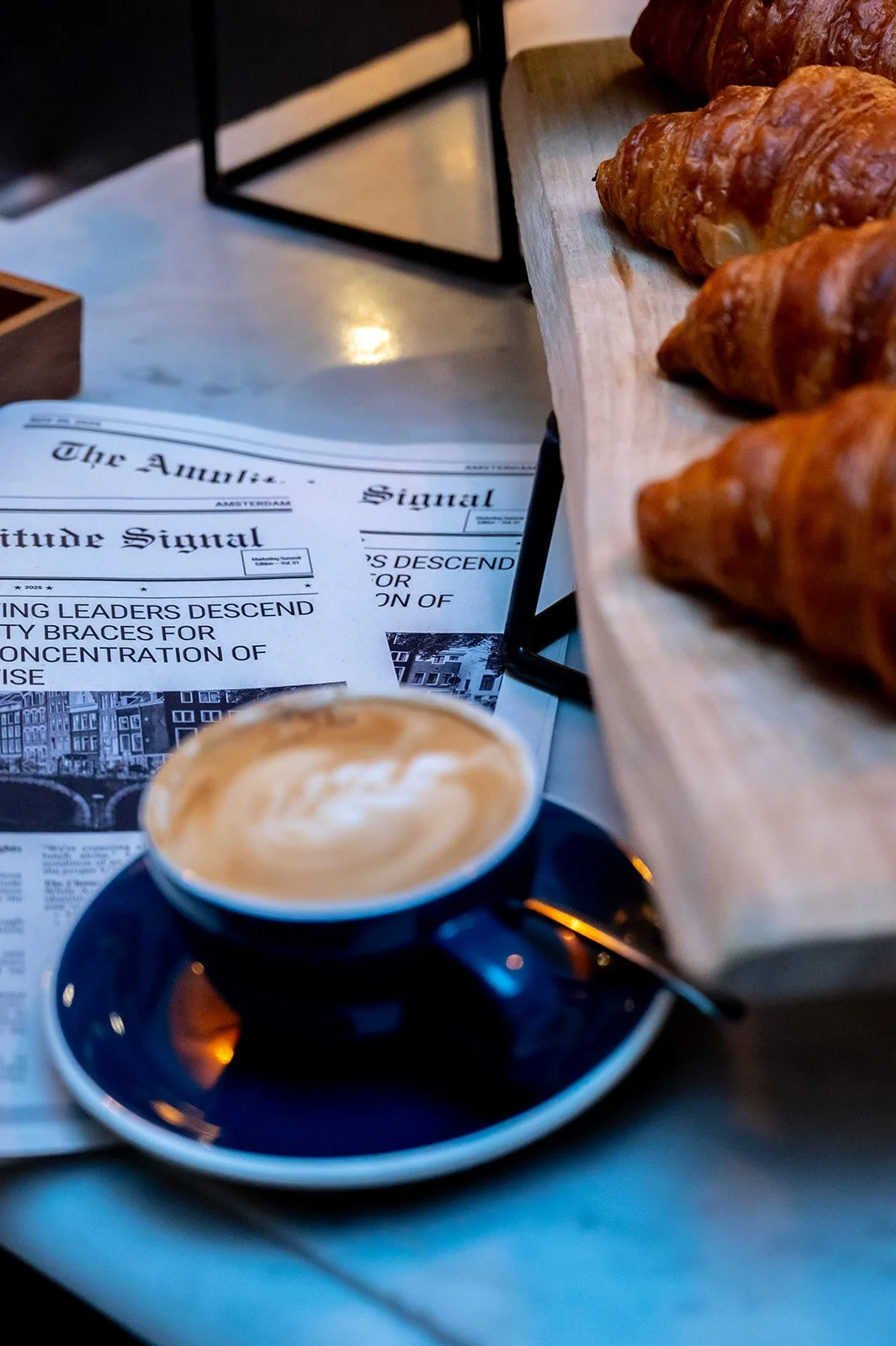 Cappuccino in een blauwe kop met schotel, croissants op een houten plank, kranten op een tafel in een koffiehuis.