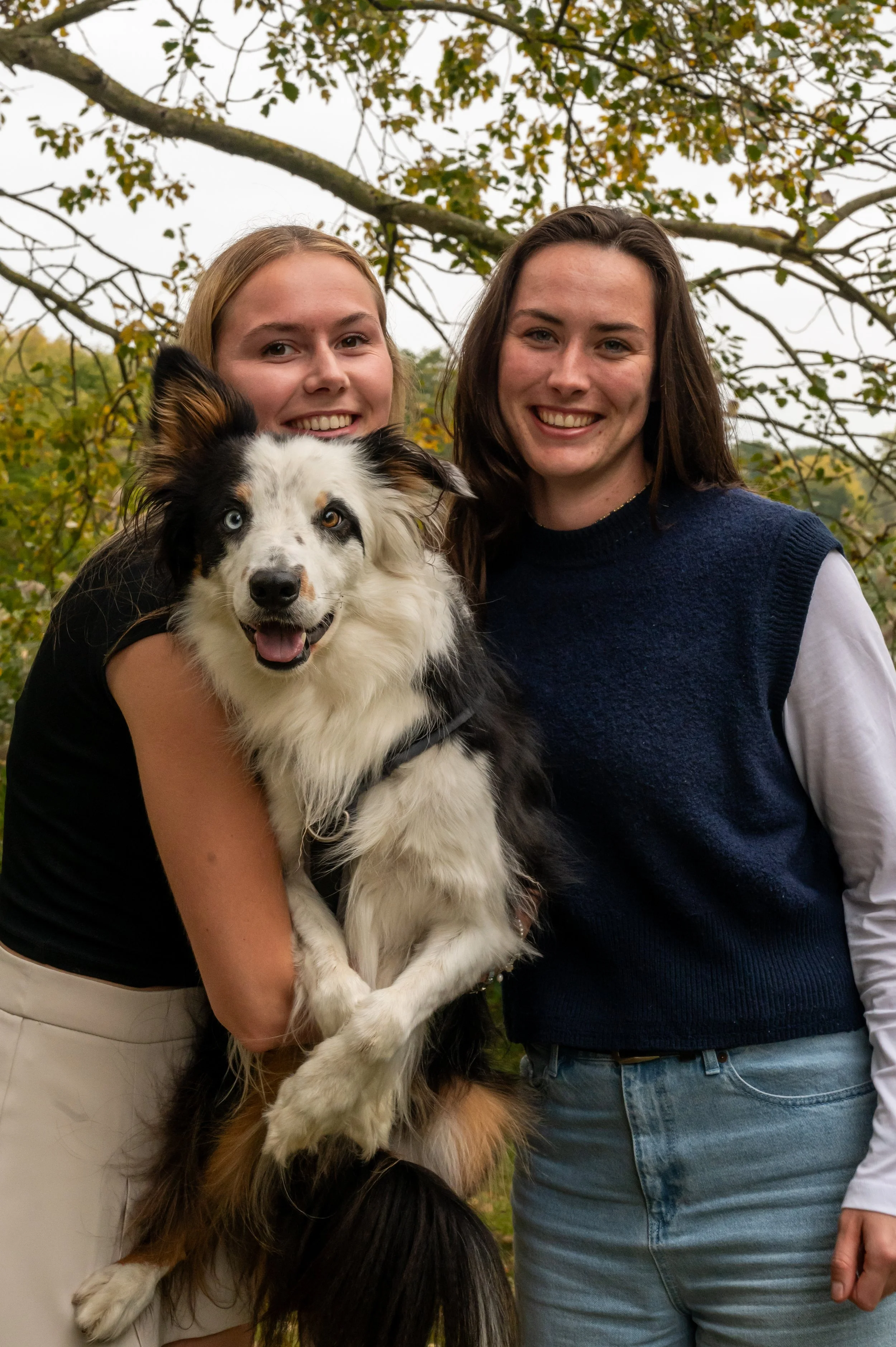 Twee jonge vrouwen met een Australian Shepherd hond buiten, met bomen en een overcastlucht op de achtergrond.