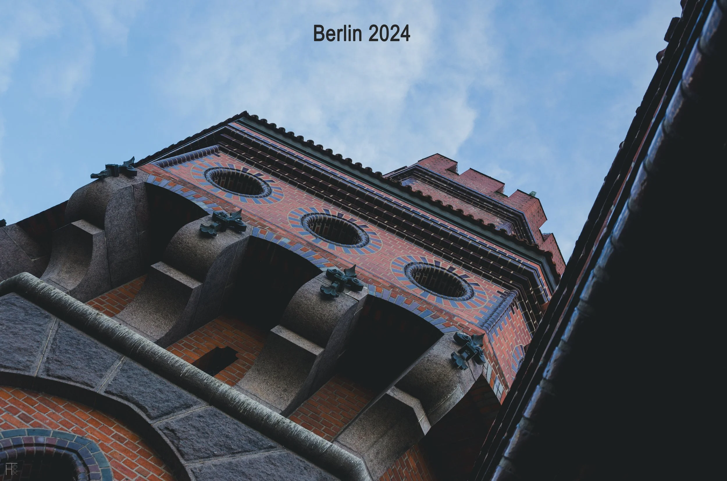 Low-angle view of a Gothic-style brick tower with circular windows against a blue sky, labeled "Berlin 2024."