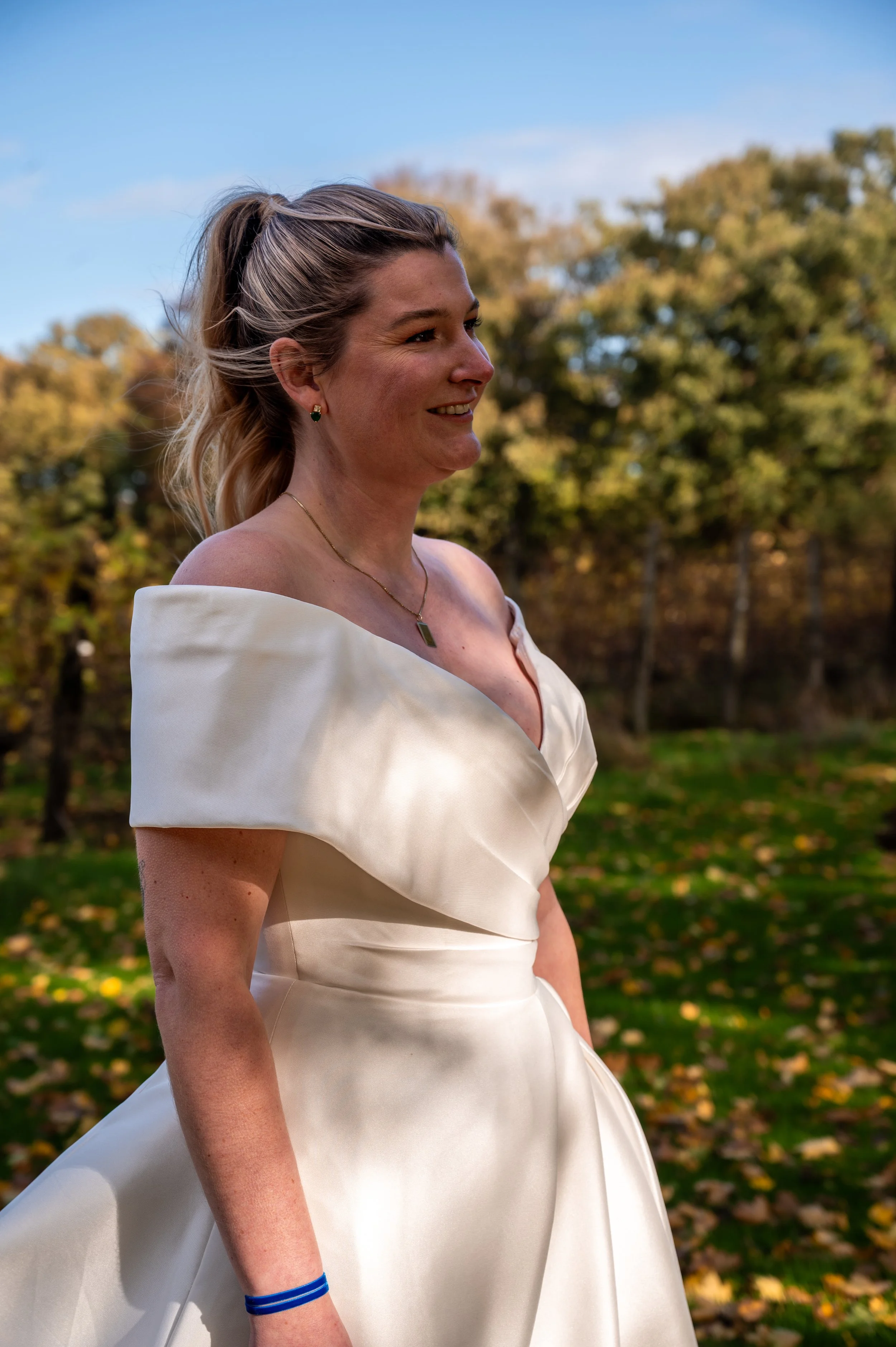 Vrouw in een witte jurk met off-shoulder ontwerp, glimlachend in een park met herfstbladeren en bomen.