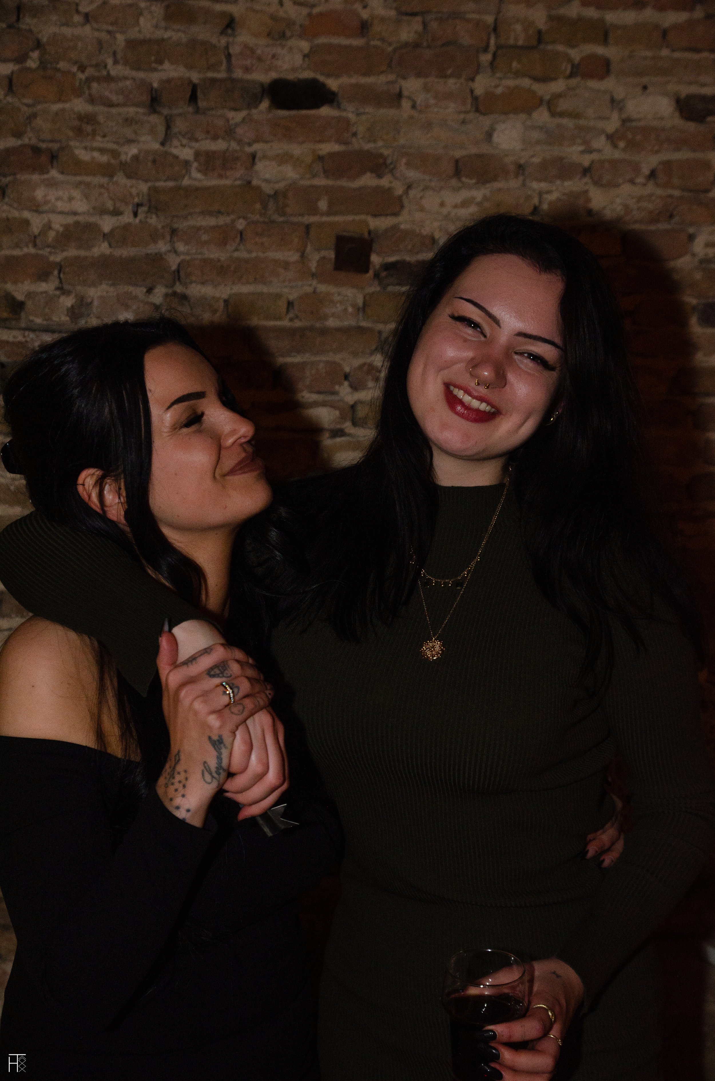 Two women smiling and embracing in front of a brick wall, with one holding a drink.