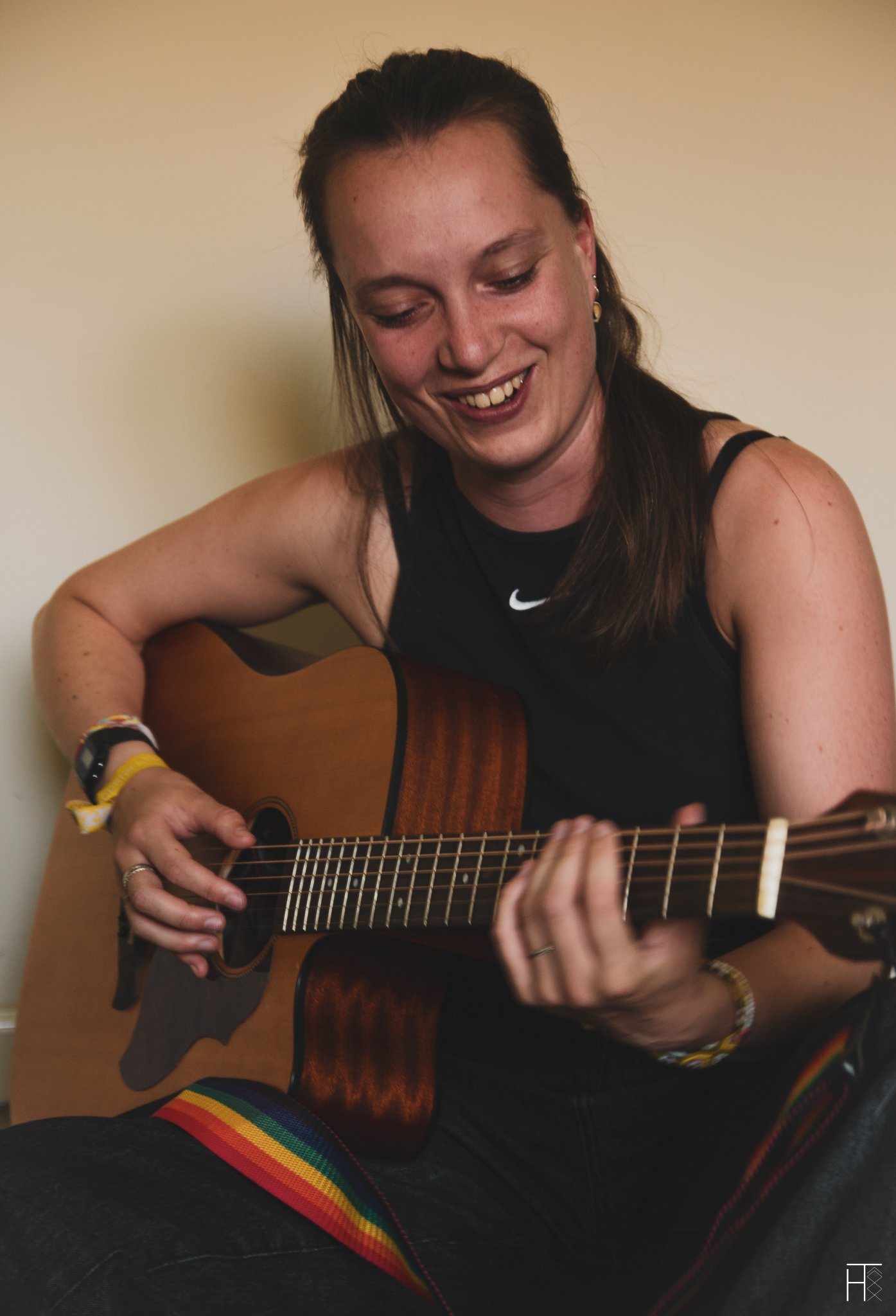 Person smiling while playing an acoustic guitar, wearing a sleeveless black top and a colorful strap.