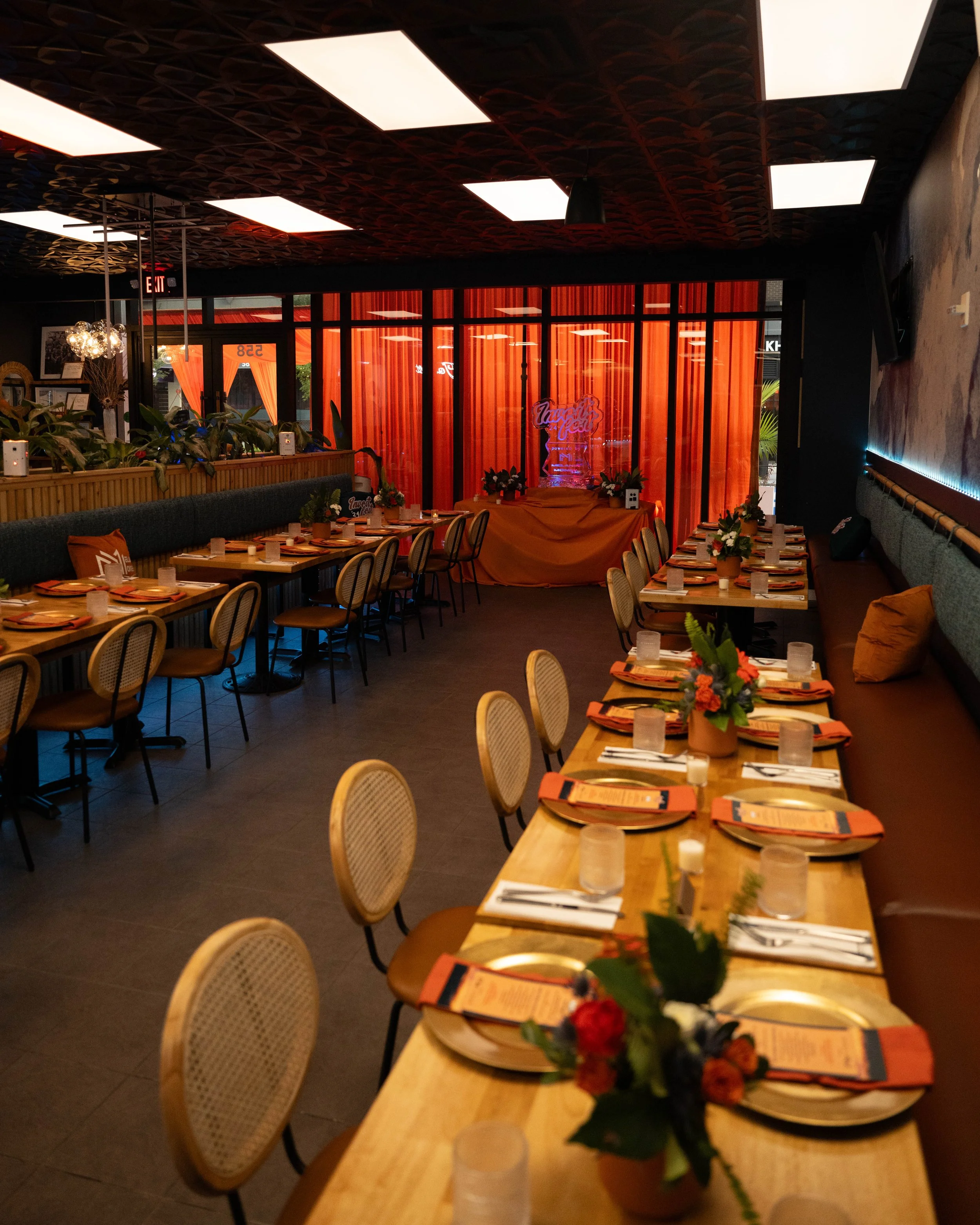 Inside a restaurant with decorated tables, orange accents, curtains, and floral centerpieces, preparing for an event or celebration.