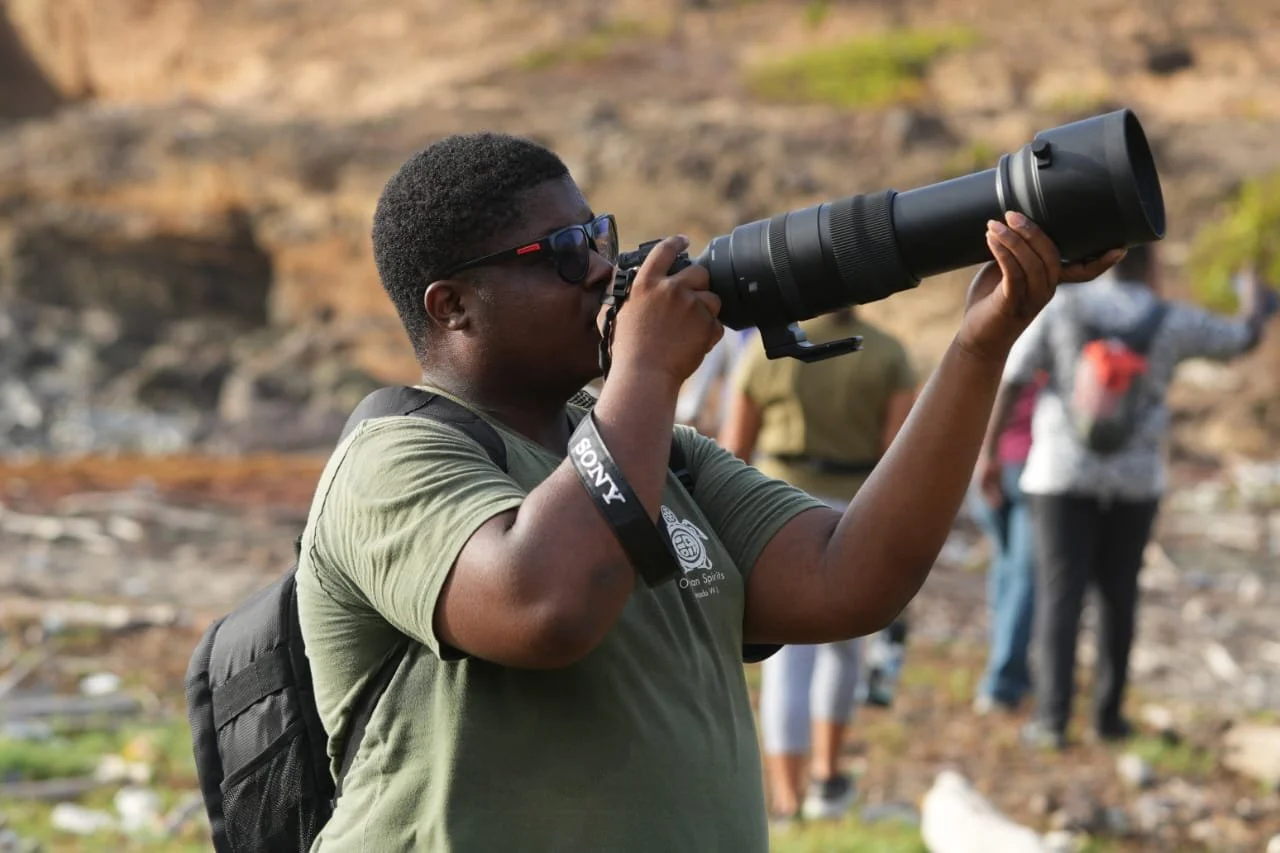 A person holding a camera with a large telephoto lens, wearing sunglasses and a backpack, outdoors with a rocky background and other people in the distance.