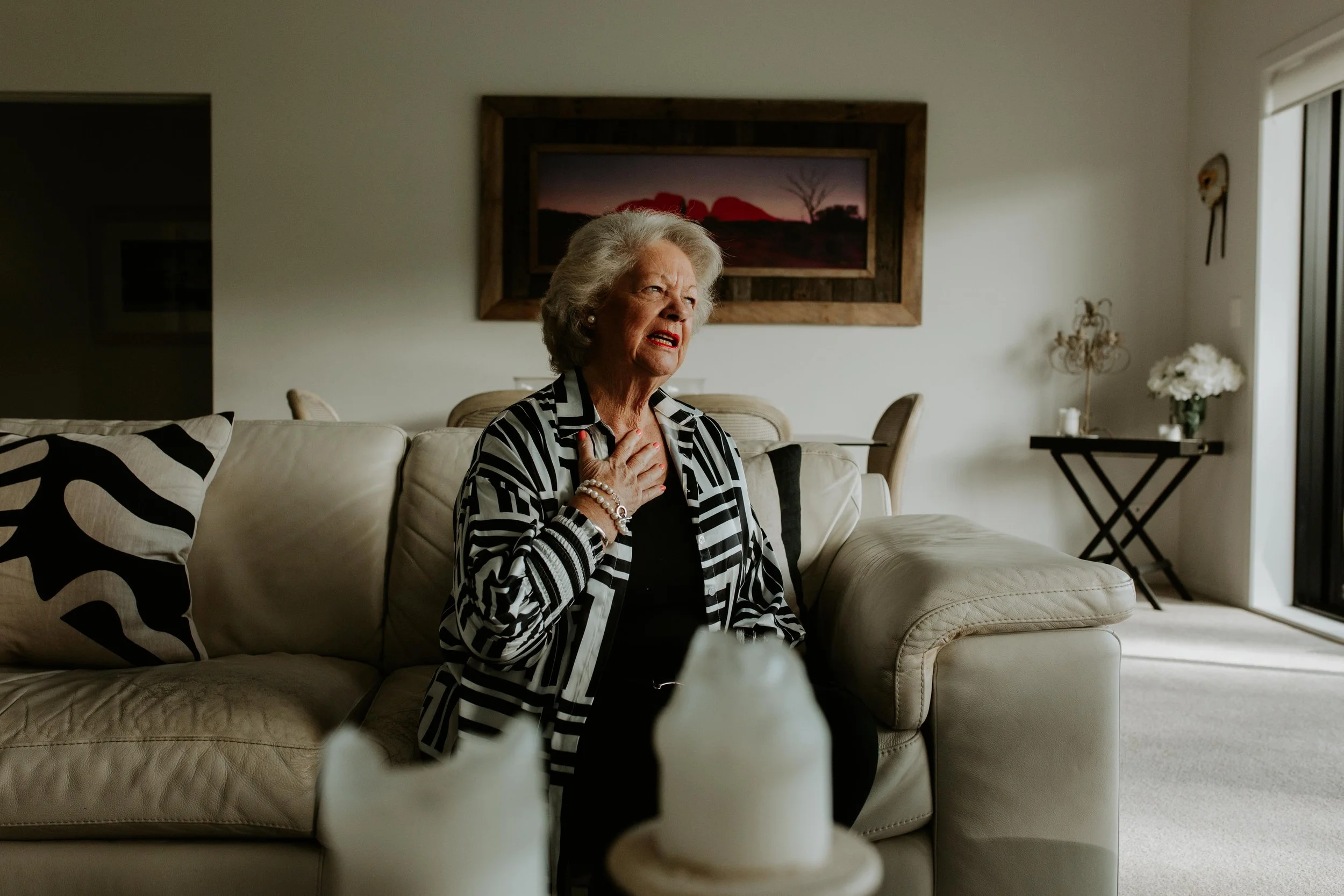 An elderly woman sitting on a beige sofa in a living room, holding her hand to her chest with a concerned expression. The room is decorated with a framed landscape painting on the wall, a small table with white flowers, and candles, with sunlight coming through a sliding glass door.