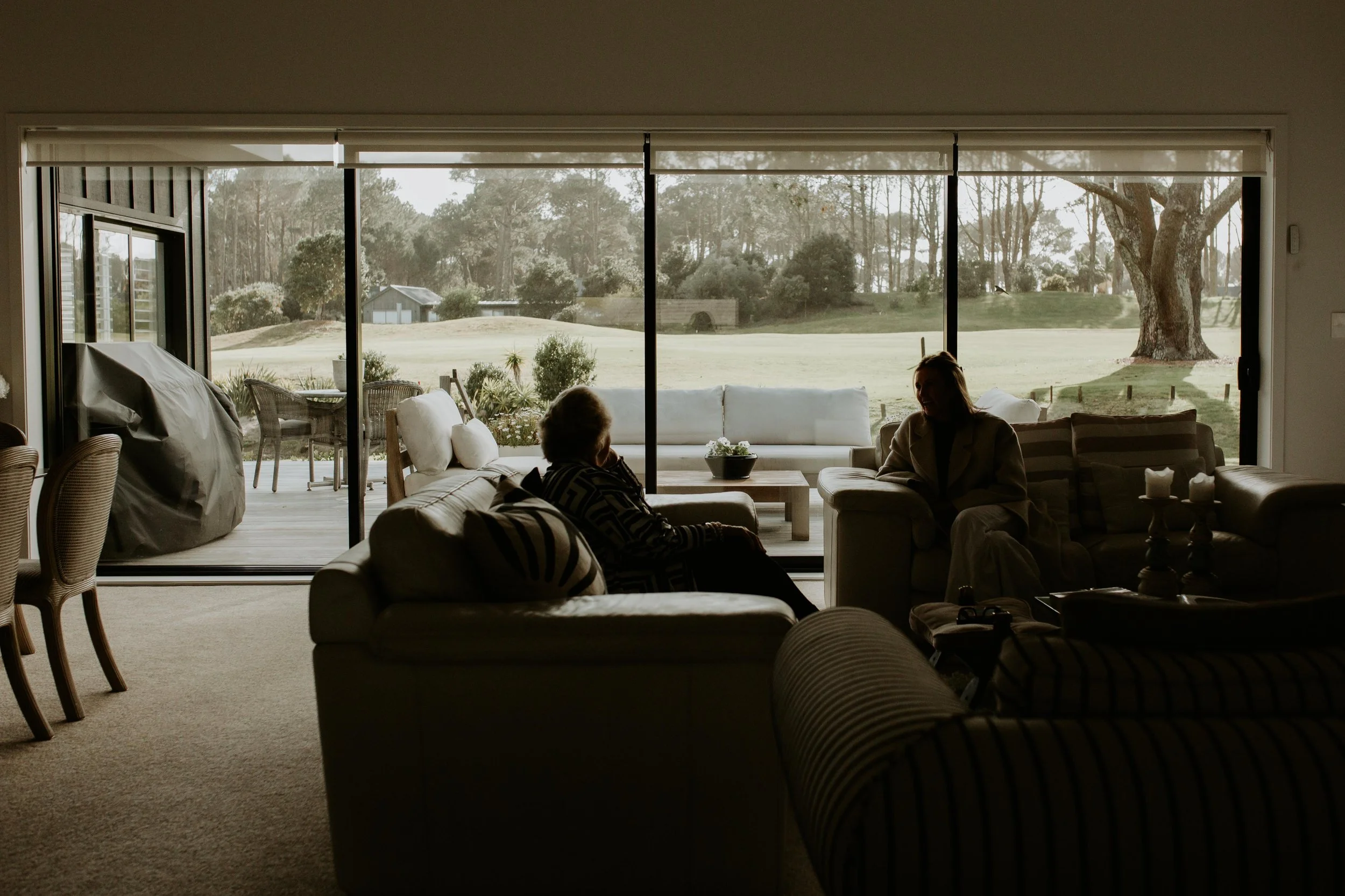Silhouettes of two women sitting in a living room, looking outside at a large yard and trees through glass sliding doors.