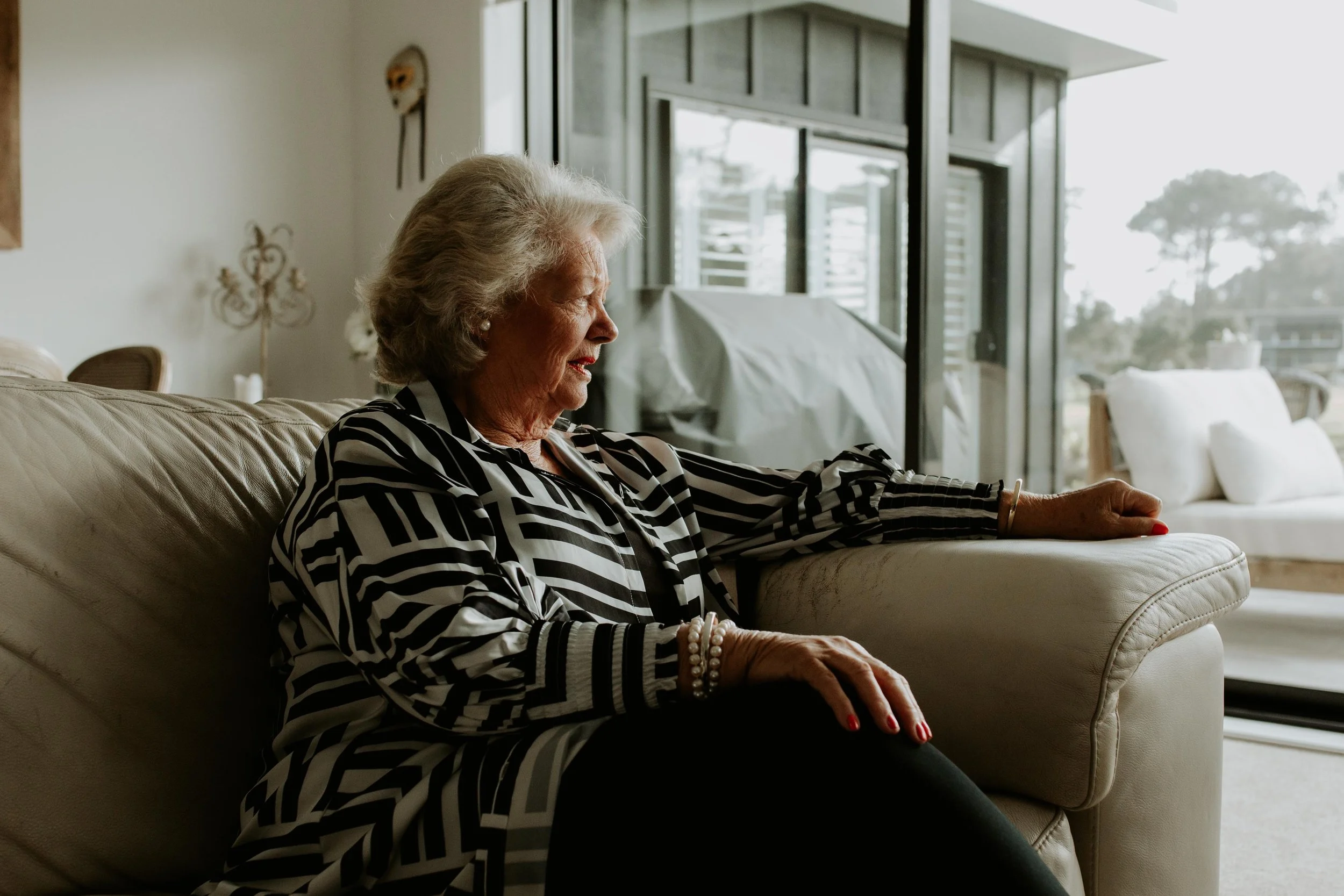 An elderly woman with gray hair sitting on a beige sofa in a modern living room, looking out a glass sliding door. Mary Sharp is holding a deck of angel cards giving a clairvoyant reading