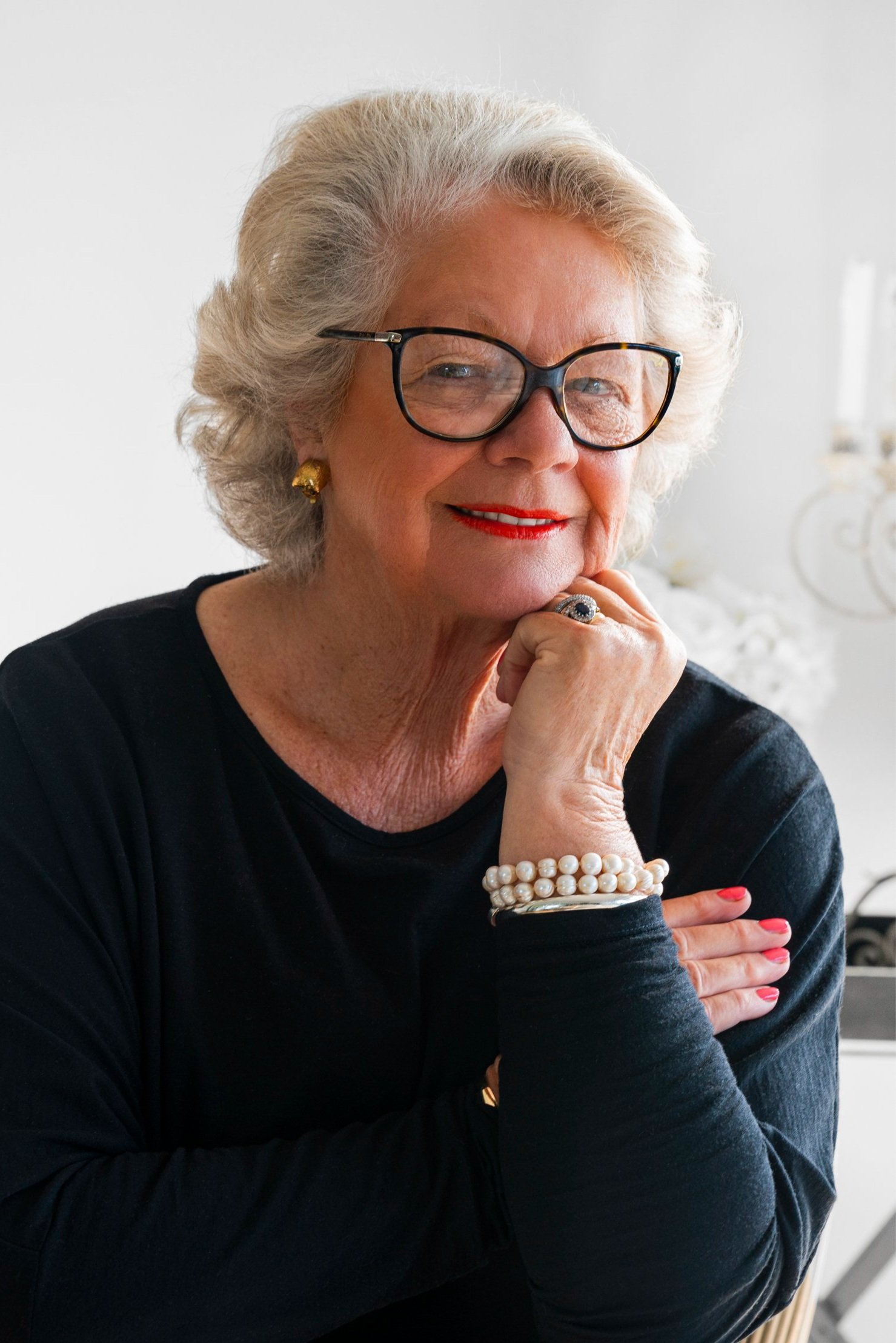 A smiling elderly woman with gray hair, wearing glasses, gold earrings, red lipstick, a pearl bracelet, and a ring, resting her chin on her hand against a bright, neutral background.