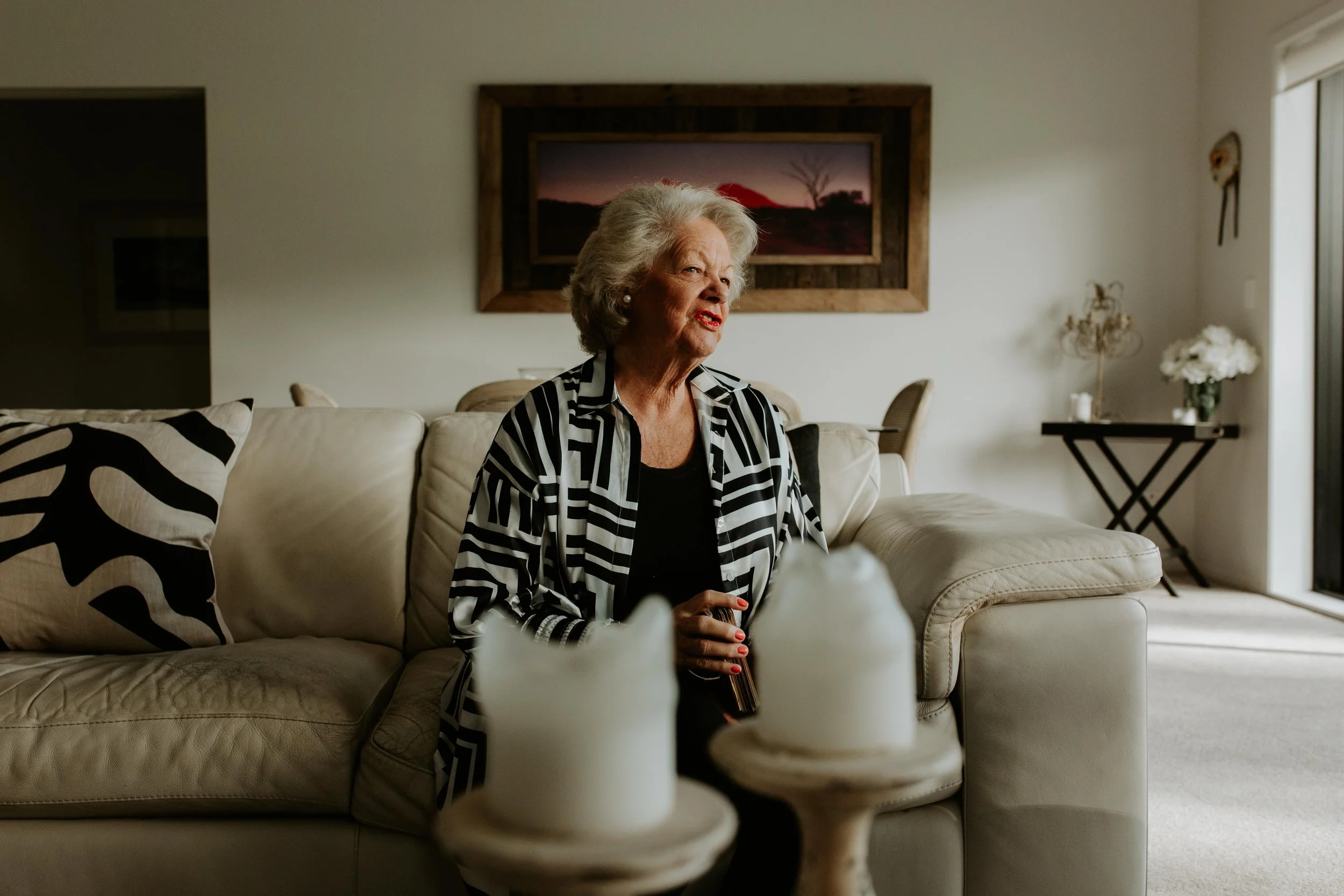 An elderly woman with gray hair, dressed in a black top and a black and white striped jacket, sitting on a white sofa in a living room, Mary Sharp is holding a deck of angel cards giving a clairvoyant reading