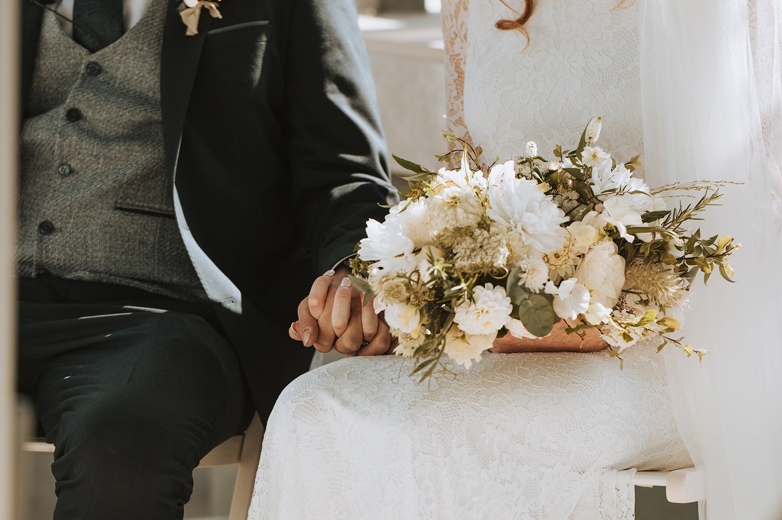 A bride sitting at a wedding ceremony holding a bouquet of white flowers, with a groom sitting beside her. The bride is wearing a white lace dress, and the groom is in a gray suit with a black coat.