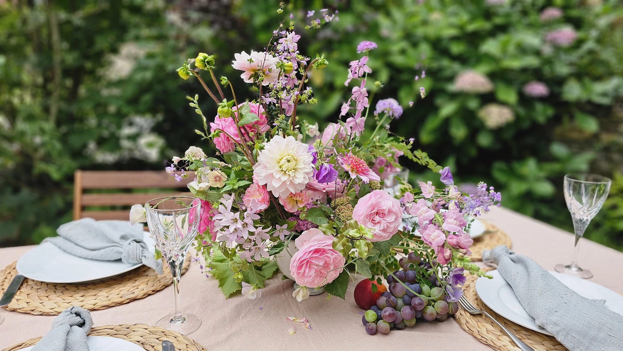 A floral centerpiece on an outdoor table set with plates, glassware, and napkins for a meal, surrounded by greenery.