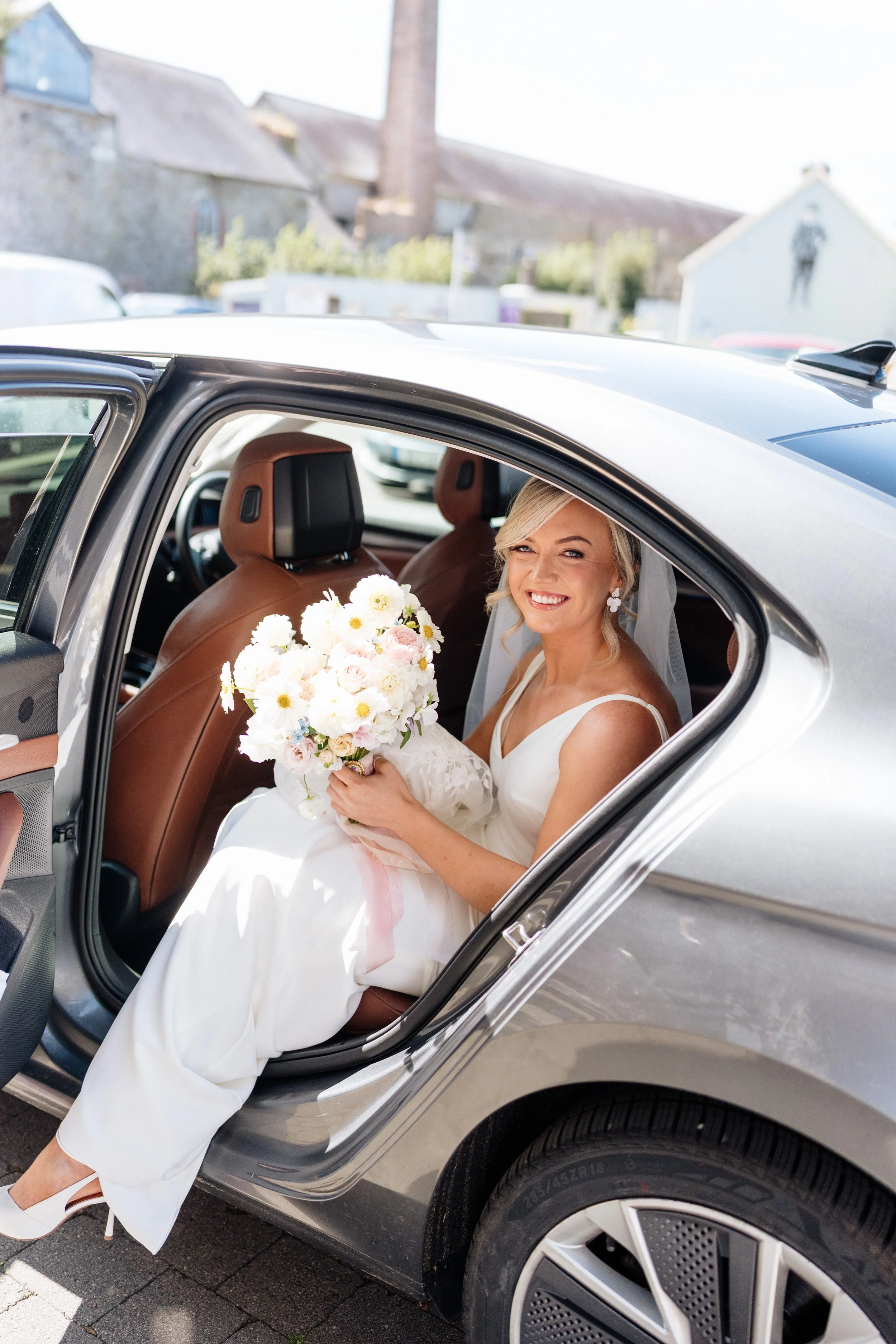 bride sitting in a car smiling holding a bouquet of white, pink and blue flowers