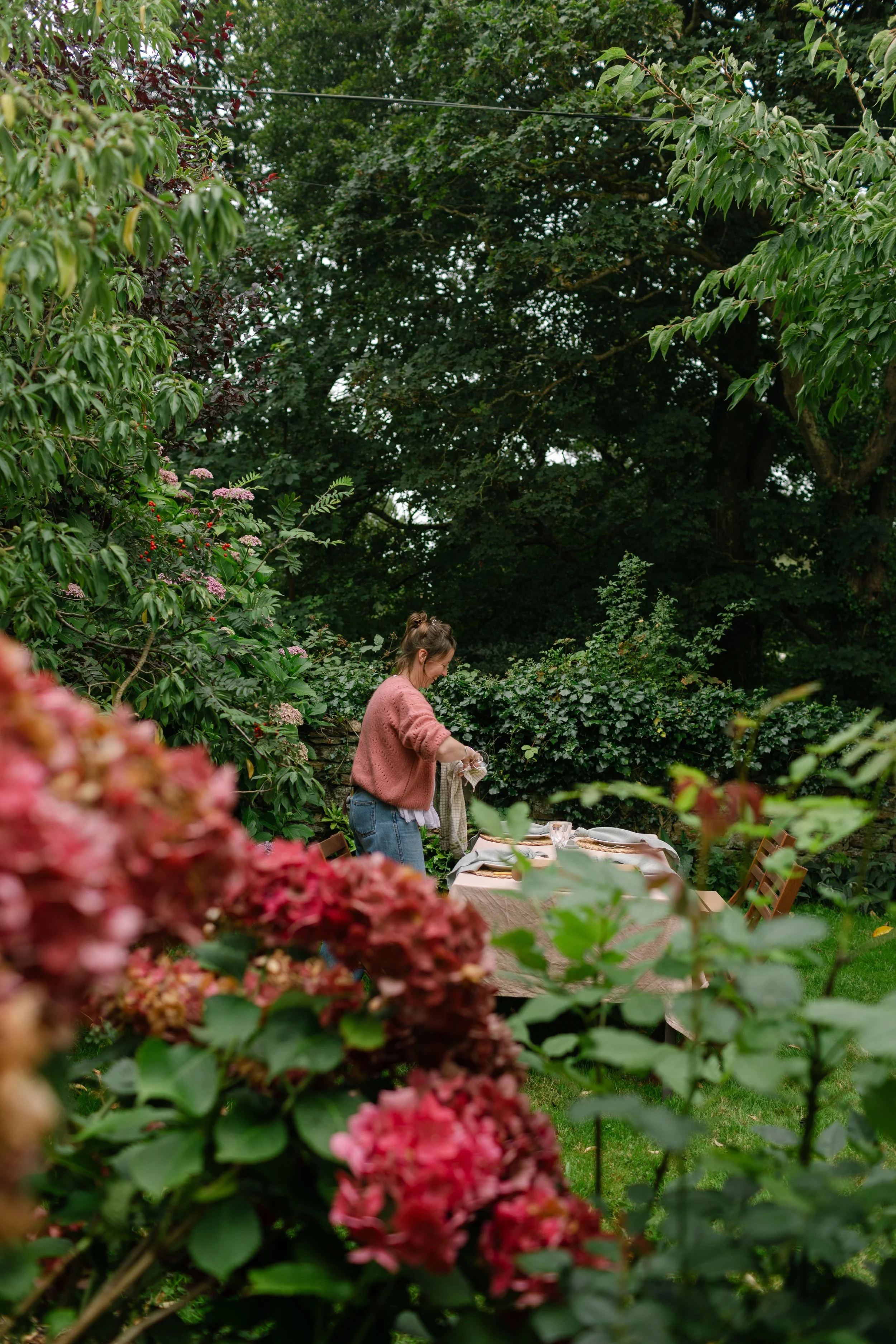 A woman setting up an outdoor dining table in a lush garden with green trees and shrubs, partially blurred pink hydrangeas in the foreground.