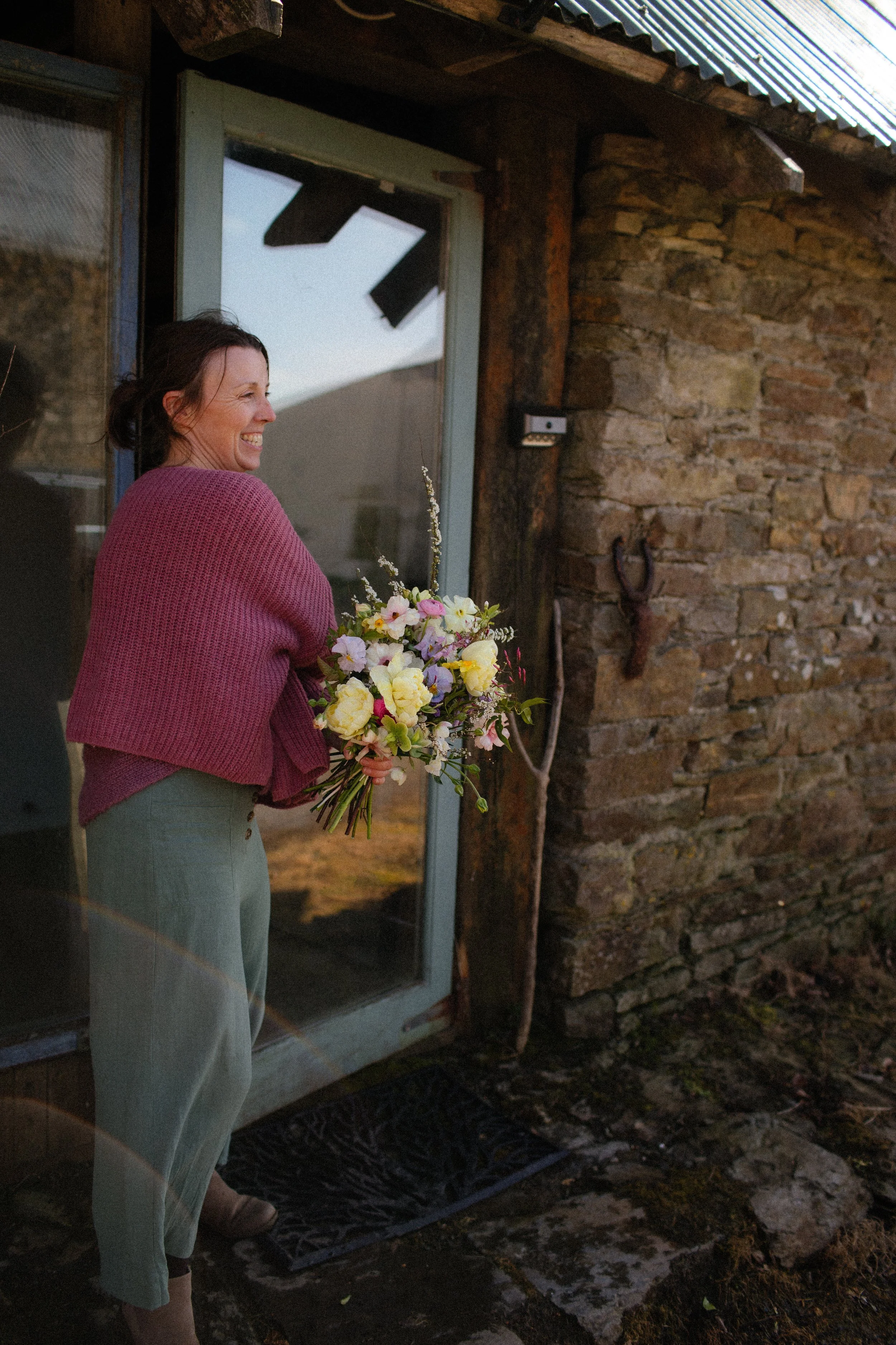 A woman wearing a pink sweater and light-colored pants holding a bouquet of flowers standing outside a rustic building with a stone wall, a glass door, and a horseshoe mounted on the wall.