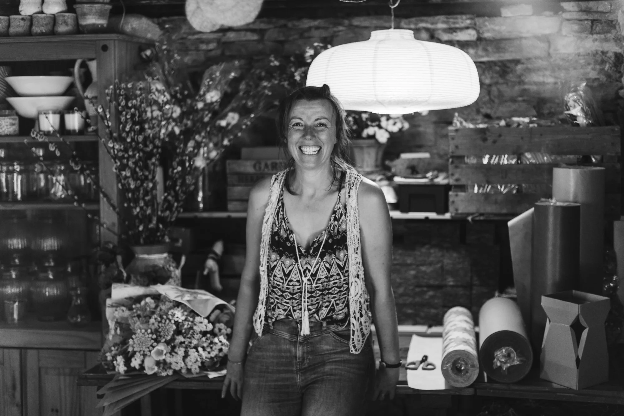 A woman smiling in a rustic indoor setting with wooden walls and shelves, a hanging paper lantern overhead, and items like rolls of wrapping paper and flowers around her.