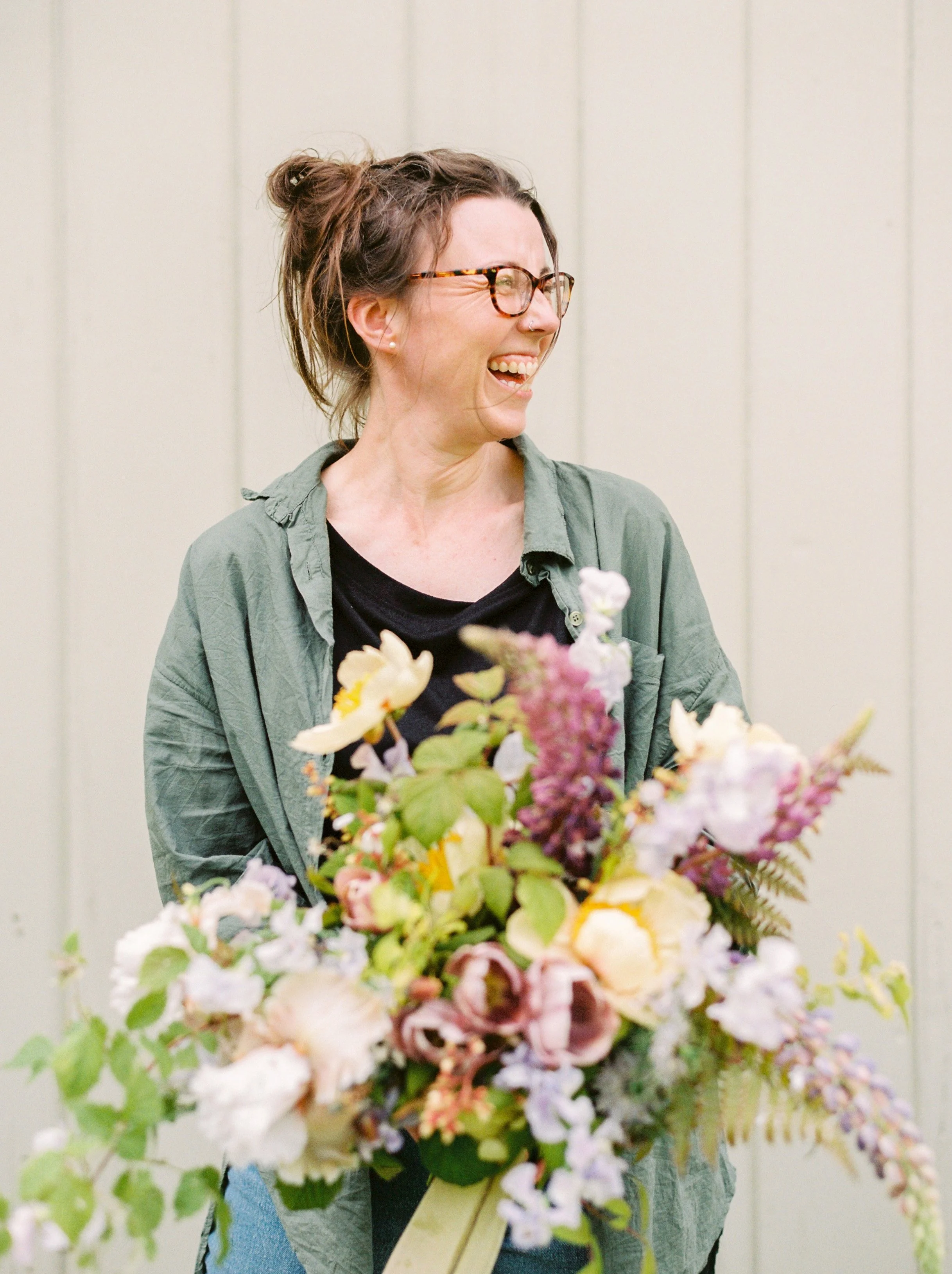 A woman with glasses and a bun hairstyle is laughing while holding a large bouquet of colorful flowers.