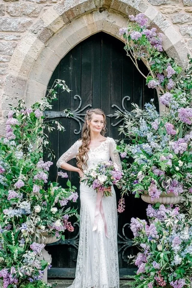 A bride in a white lace wedding dress holding a bouquet of purple and white flowers, standing in front of a black arched door surrounded by lush purple and white flowers and greenery.