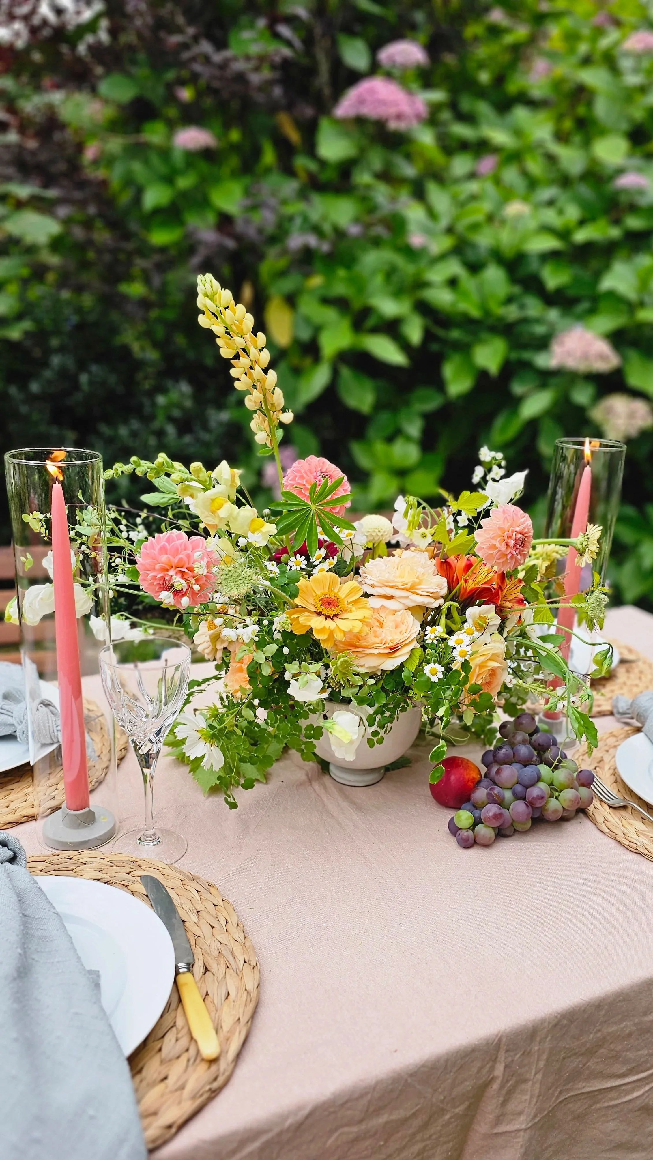 A table decorated with a large floral centerpiece, pink candles in tall glass holders, grapes, an apple, and a white plate with a folded napkin and a yellow utensil, set outdoors with greenery in the background.