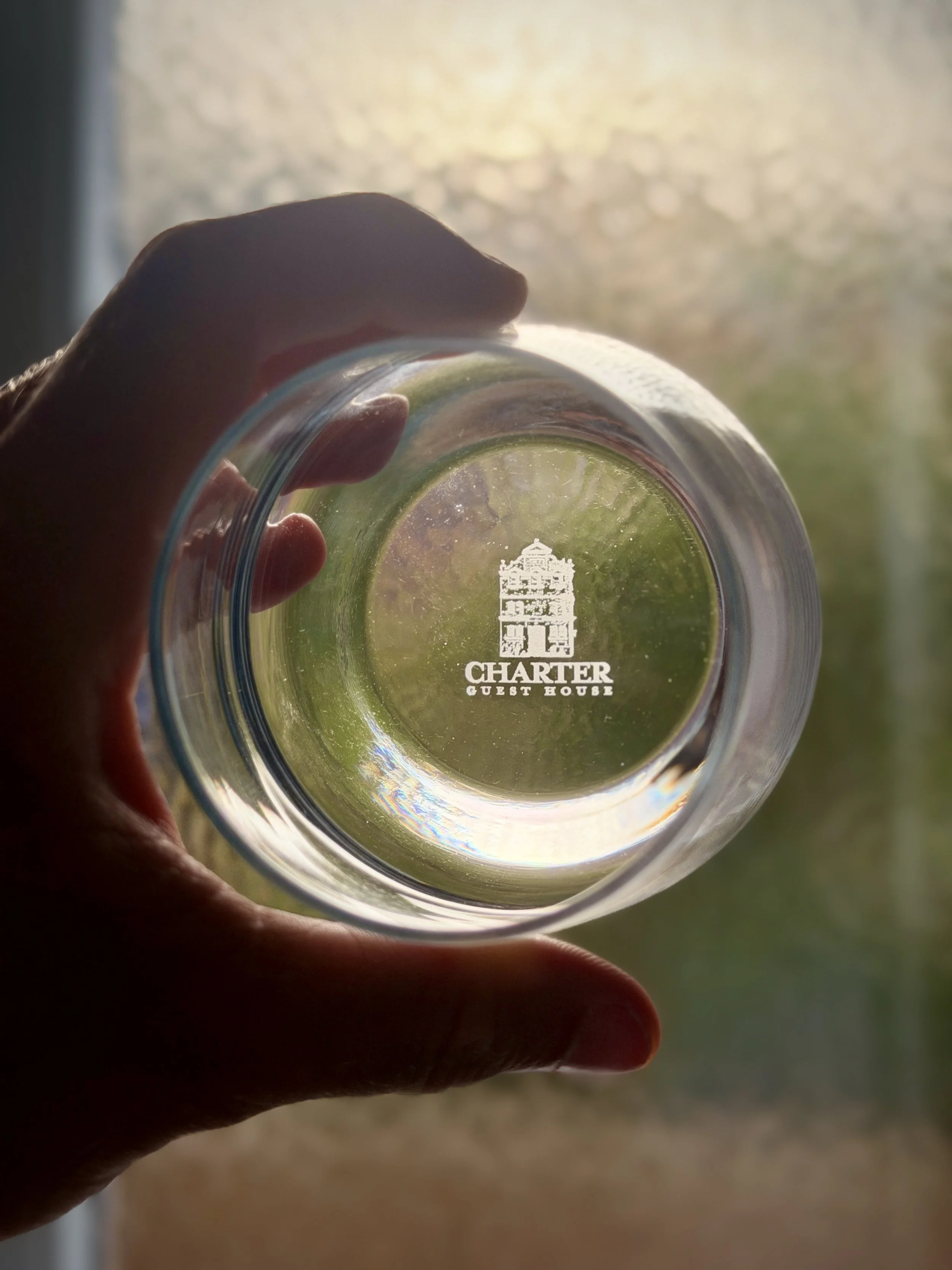 A hand holding a clear glass with the 'Chatter Guest House' logo, viewed through the glass. The background is blurry and sunlit.