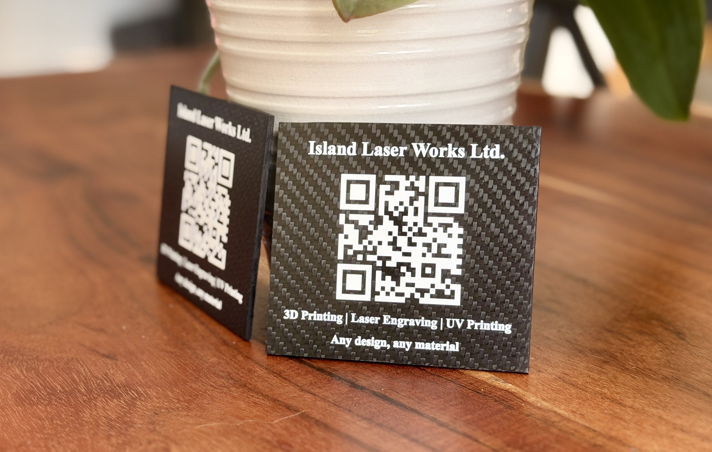 Close-up of a business card with a QR code, from Island Laser Works Ltd., placed on a wooden surface with a white ceramic pot and a green plant in the background.