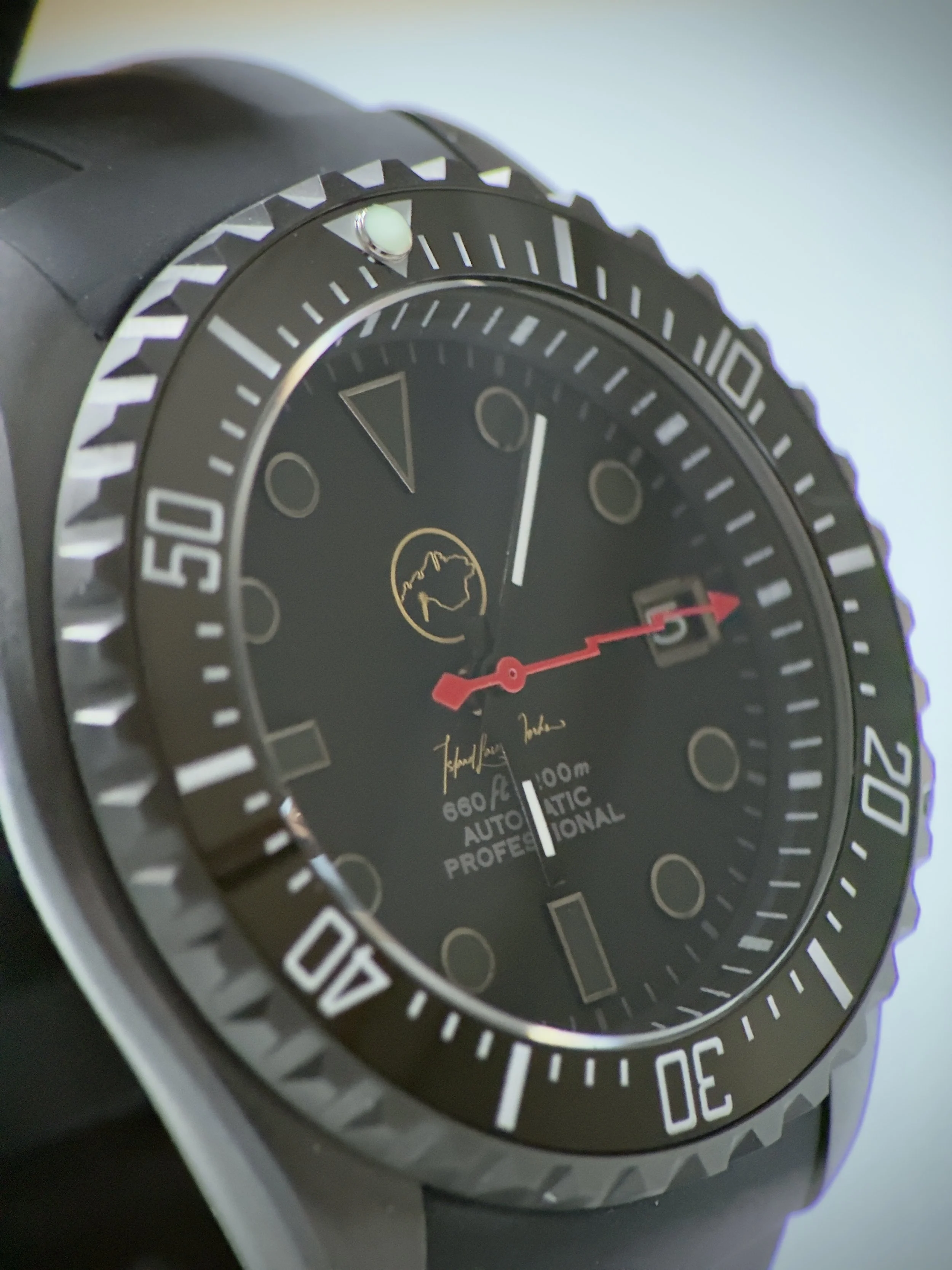 Close-up of a black wristwatch with a rotating bezel, black dial, and red seconds hand, featuring a gold outline map of Australia logo and some signature text on the dial.