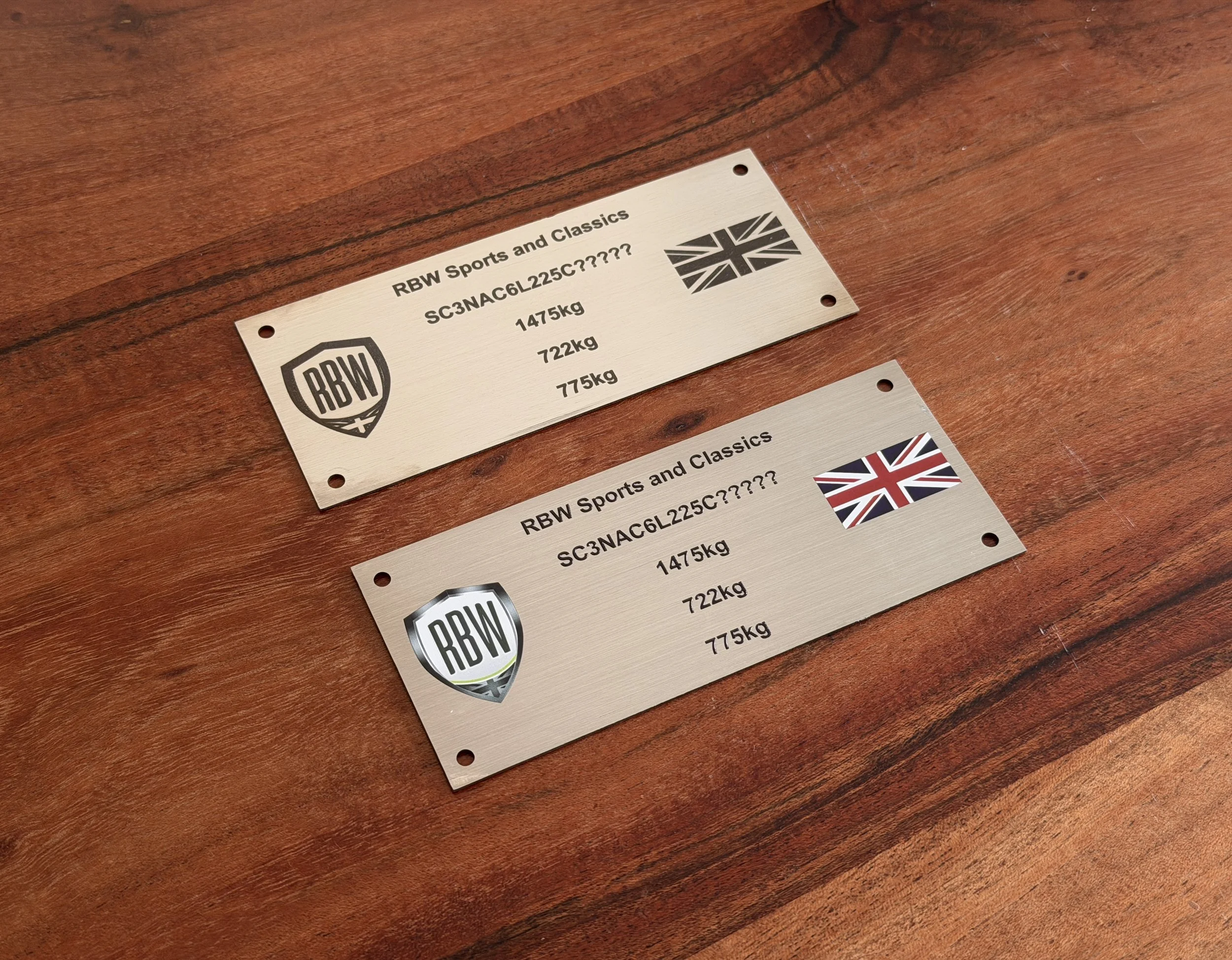 Two metal nameplates with black text and logos, placed on a wooden surface, one with a UK flag and the other with a Union Jack flag.