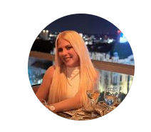 A woman with long blonde hair smiling at a dinner table with a cityscape in the background at night