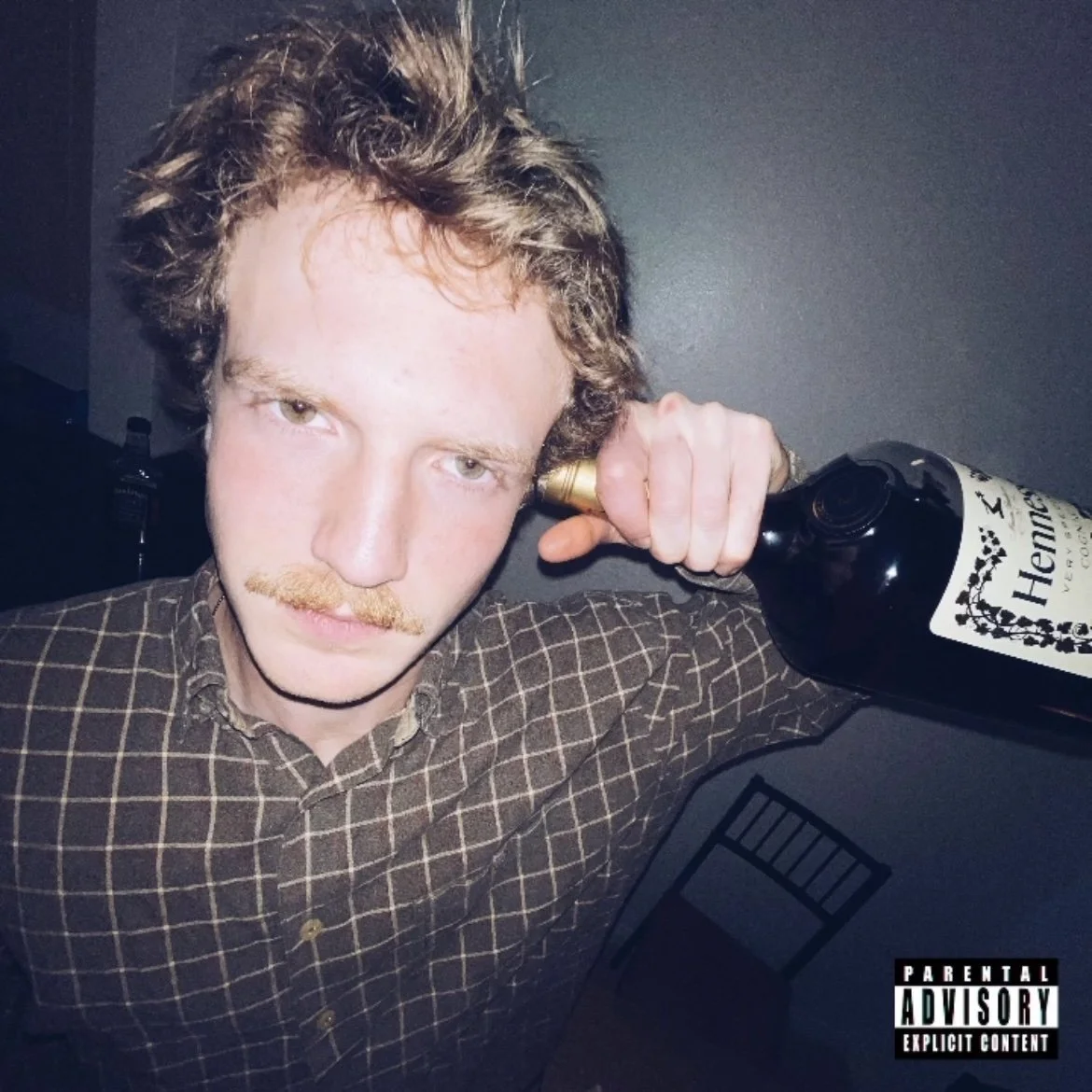 Christopher Clucas holding a bottle of Hennessy to his ear, with a serious expression, wearing a brown checkered shirt, in a dimly lit room.