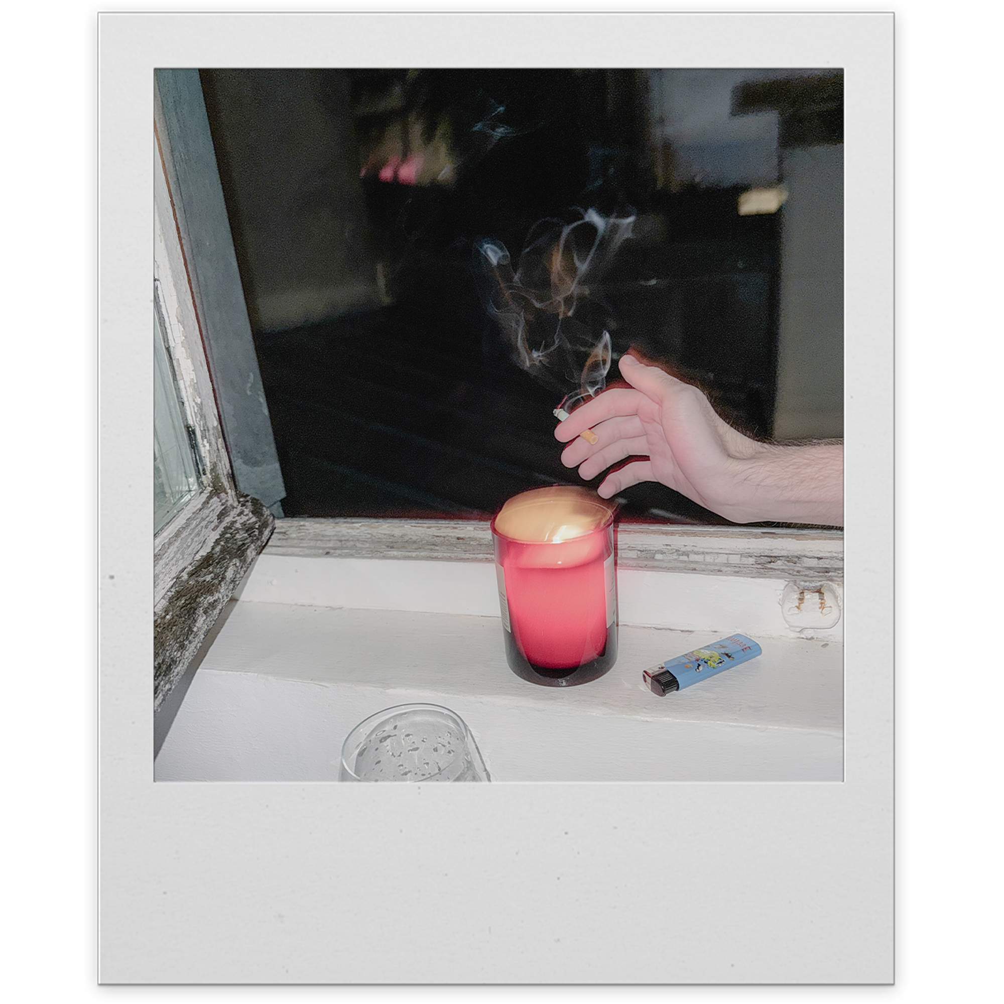 Person lighting a cigarette with a lighter near a window, with a candle and a pack of cigarettes visible on a windowsill.