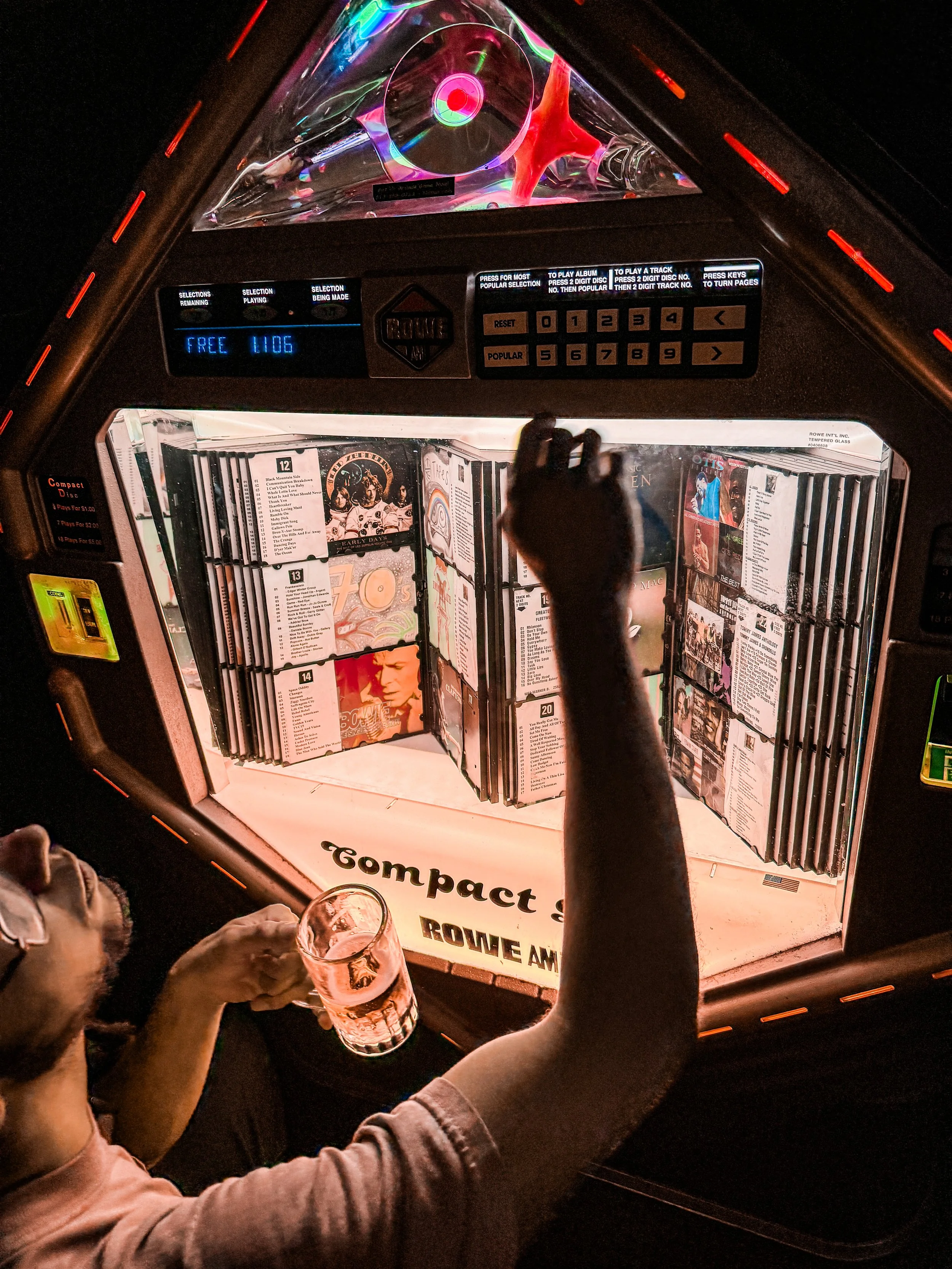A person holding a glass of beer in front of a vintage jukebox at a bar or restaurant. The jukebox has multiple rows of 45 RPM records inside.