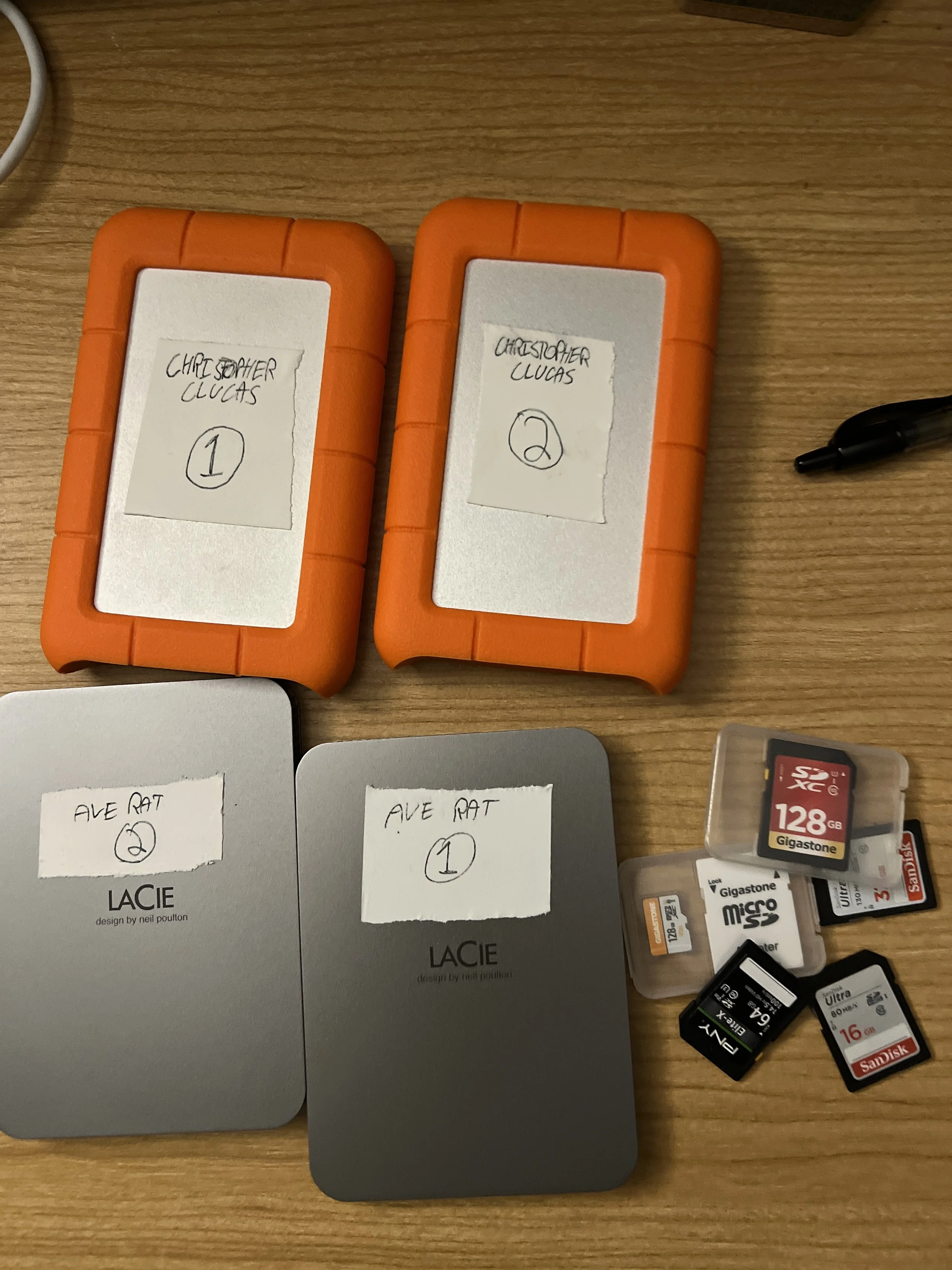 Two orange LaCie external hard drives labeled with numbers, two gray LaCie hard drives labeled similarly, and several SD and microSD memory cards scattered on a wooden surface.
