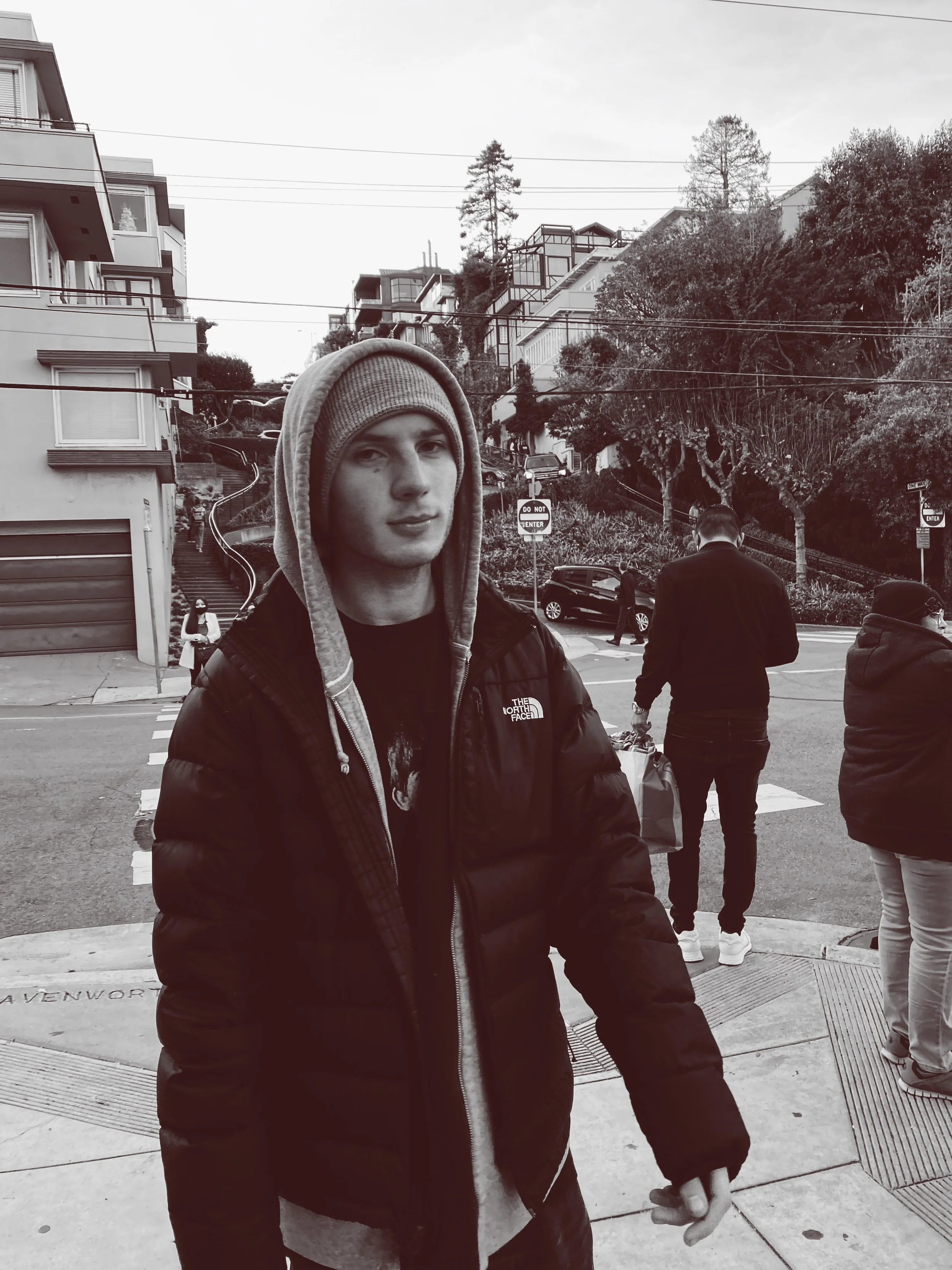 A young man in a hoodie and jacket standing on a city street, with other people, parked cars, trees, and residential buildings in the background.