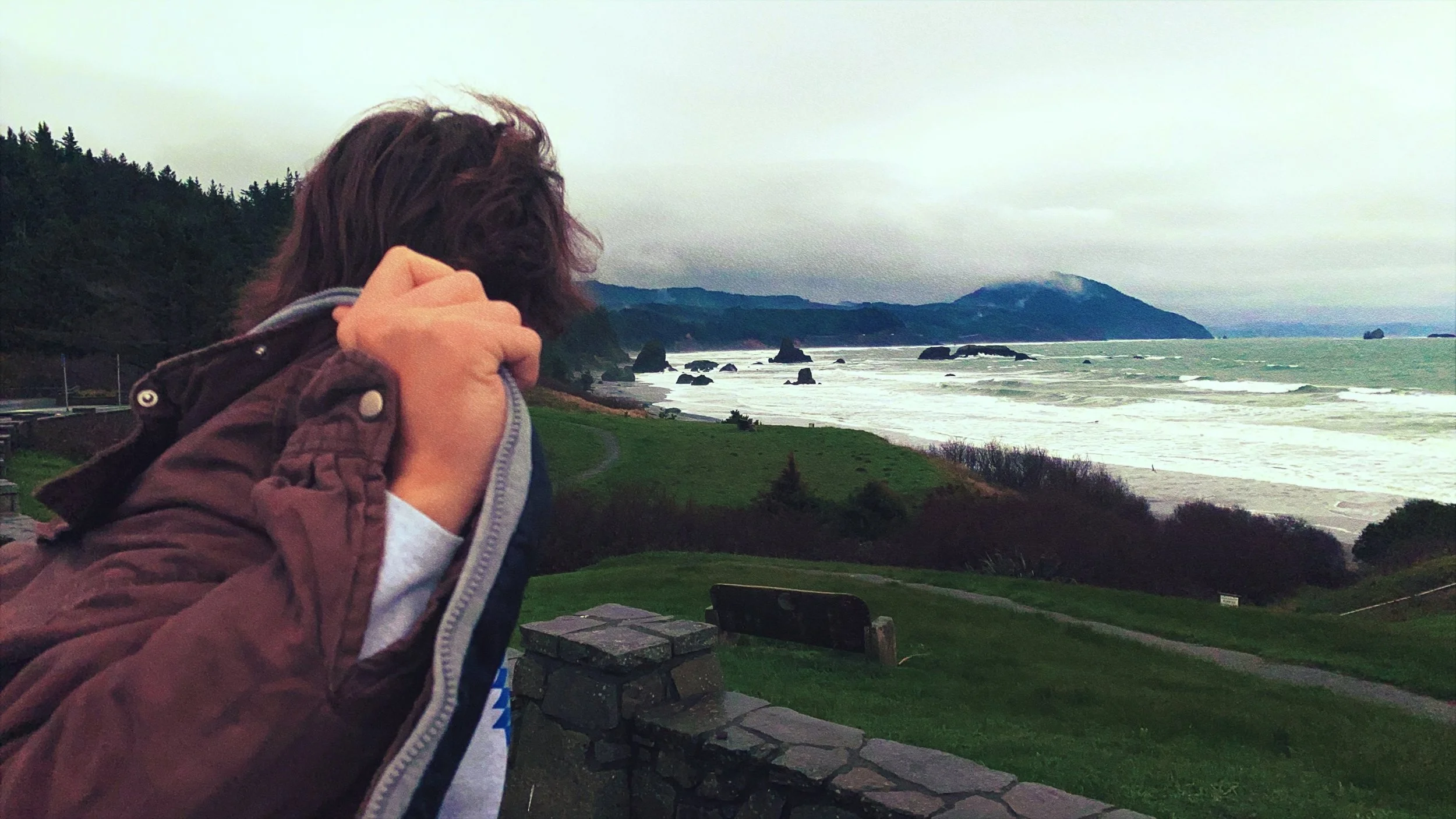 Person holding a jacket near seashells and rocks at a coastal landscape with shoreline, waves, and distant mountains under a cloudy sky.