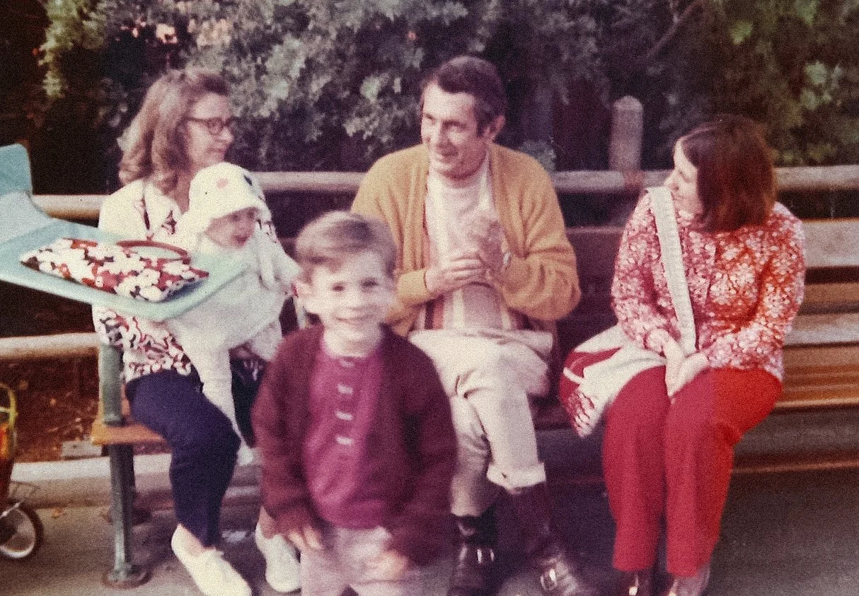 Group of five people, including three children and two adults, sitting on a park bench outdoors, engaged in conversation and smiling, with trees in the background.