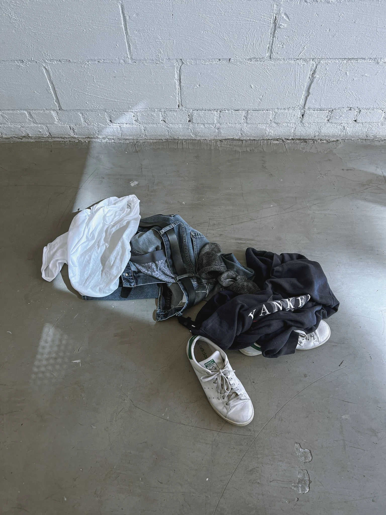 Pile of clothes including a white shirt, jeans, sneakers, and a black hoodie on a concrete floor.