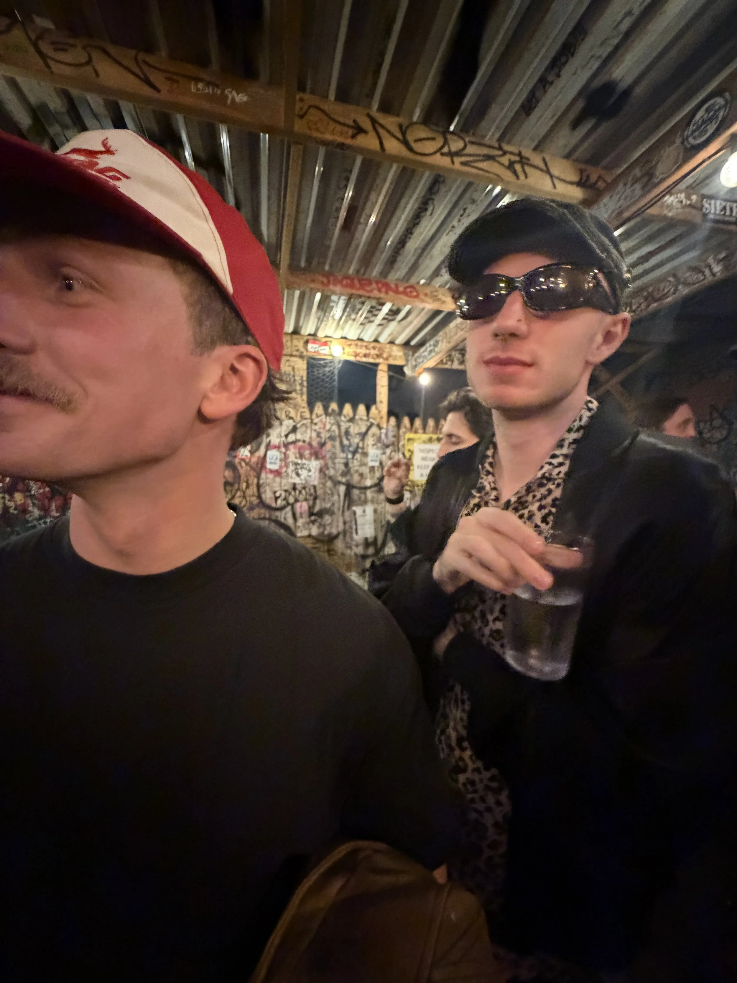 Two men inside a dimly lit bar or club with graffiti on the wood-paneled ceiling. One man is wearing a red and white cap, and the other is wearing dark sunglasses and a leopard print shirt, holding a drink.