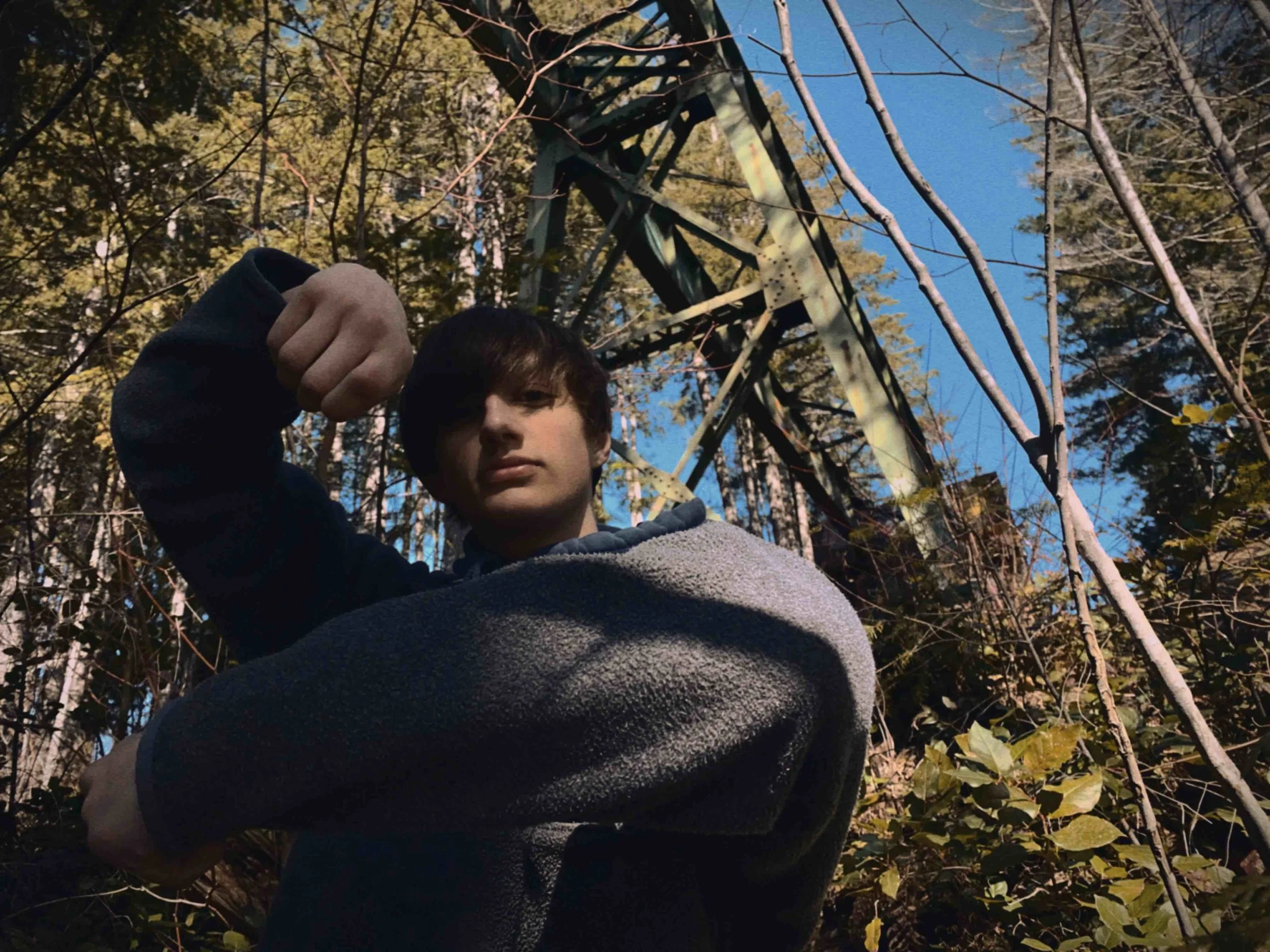 Person in a gray sweater posing in a wooded area with trees and metal structure in the background.