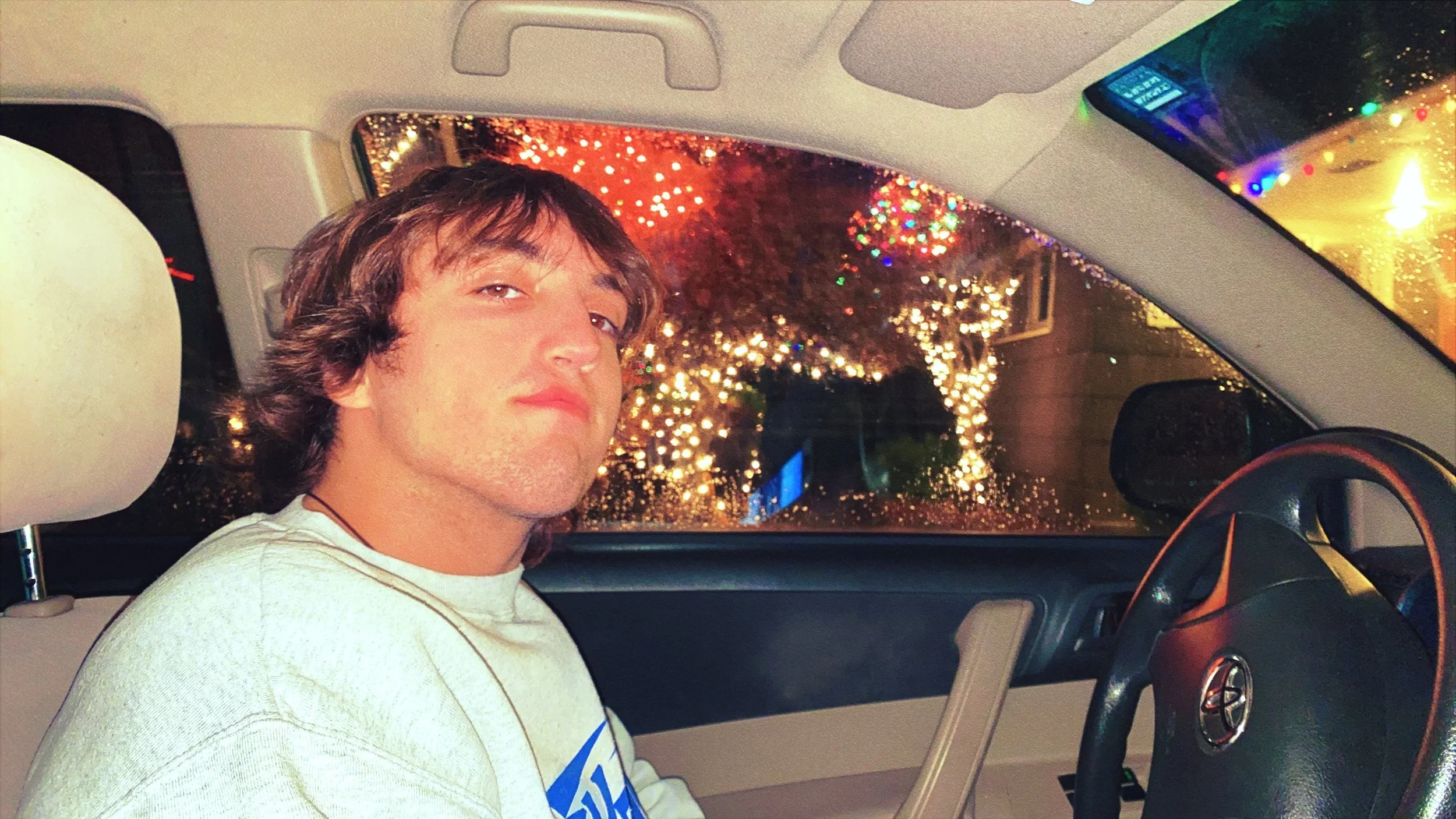 Person with dark hair looking at the camera inside a car parked during a rainy night, with water droplets on the windshield and colorful holiday lights in the background.