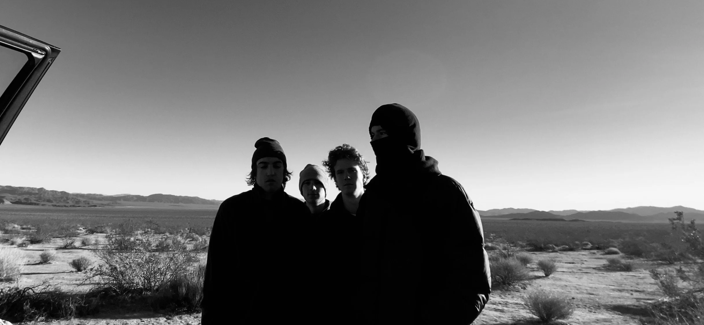 Christopher Clucas - Four young men standing outdoors in a desert landscape, wearing dark clothing and beanies, with mountains in the background and a clear sky, in black and white.