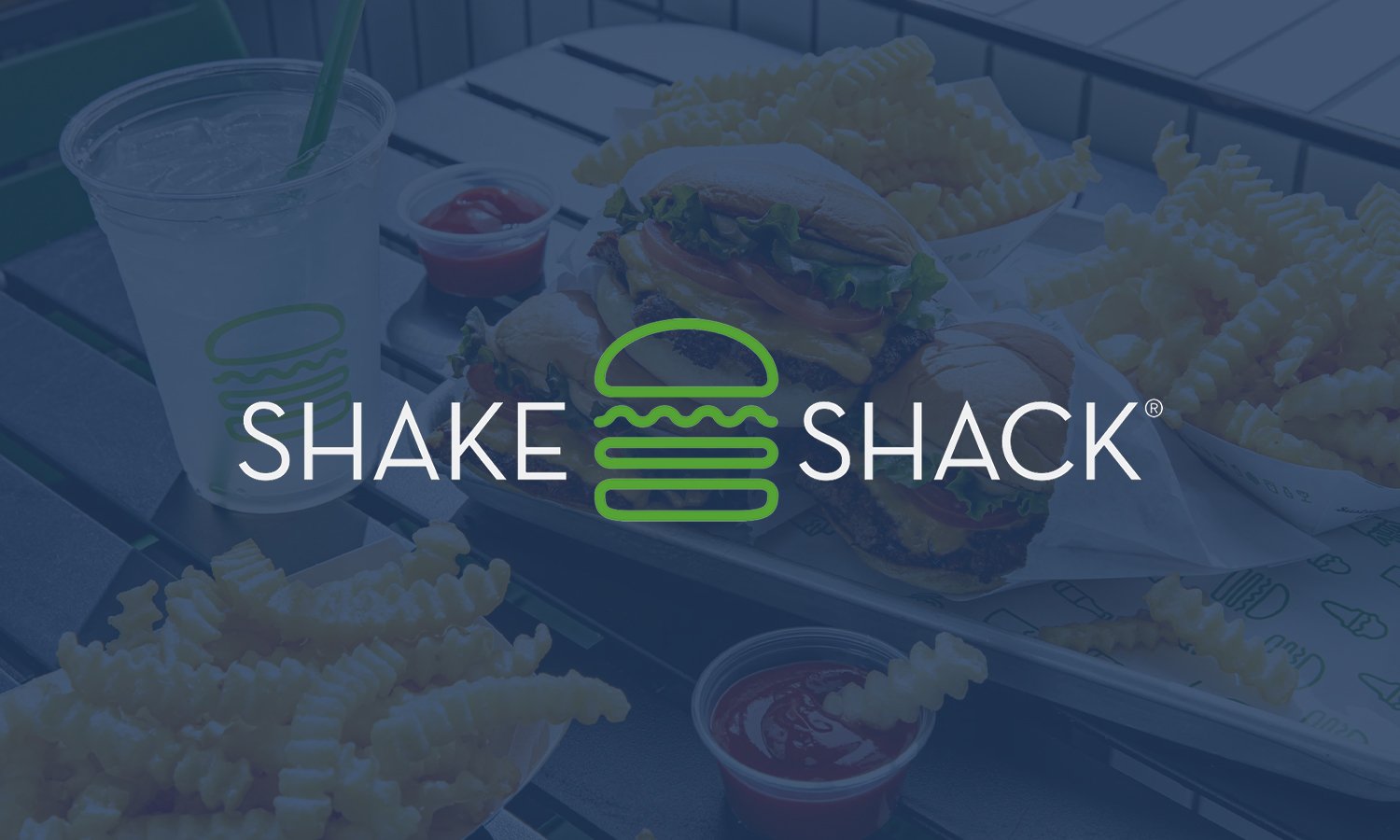 A Shake Shack meal featuring a cheeseburger with lettuce, tomato, and cheese, crinkle-cut French fries, a soft drink with a green straw, and small containers of ketchup and mayonnaise.