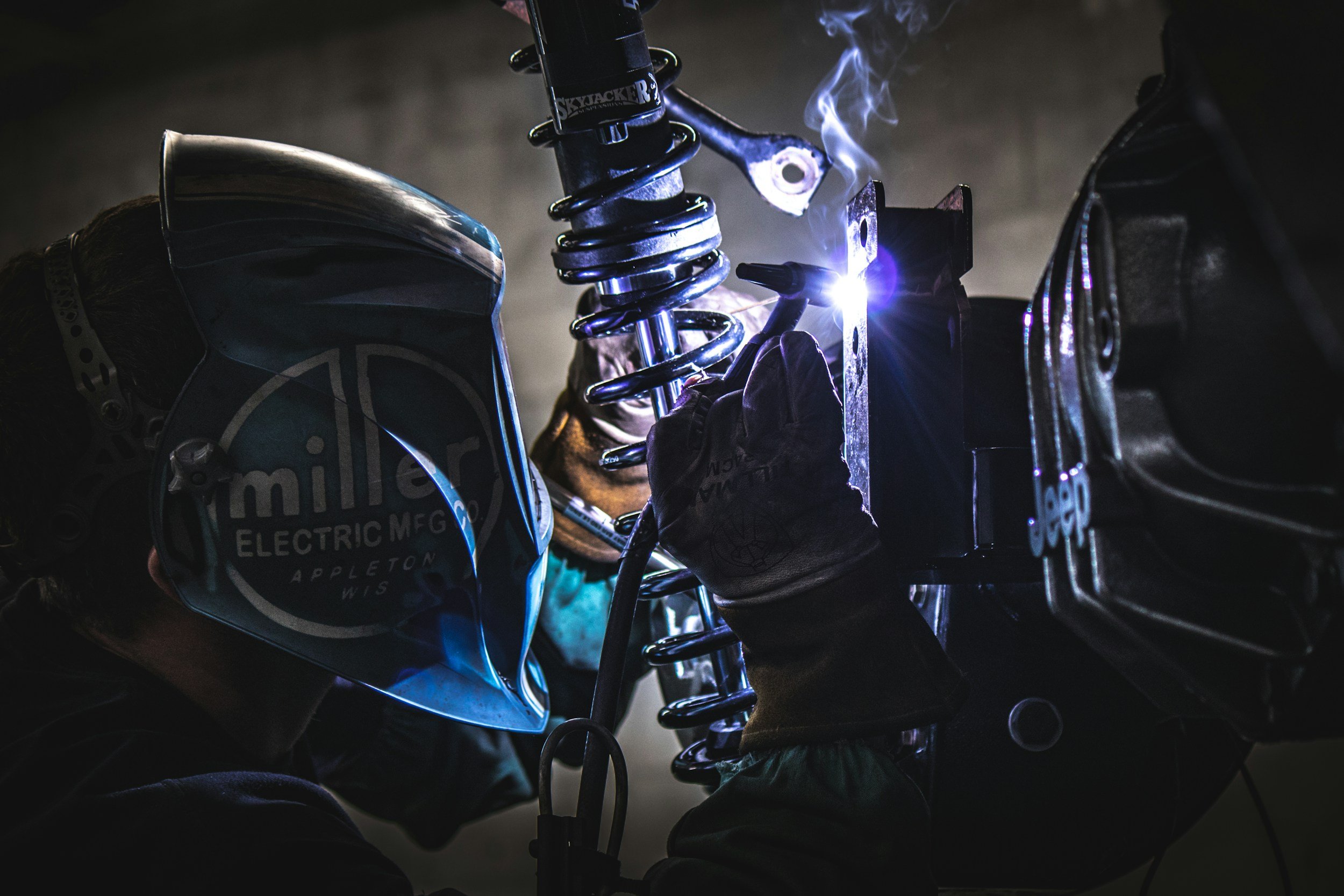 A person welding a metal part on a motorcycle suspension component, wearing a welding helmet and gloves in a dark workshop.