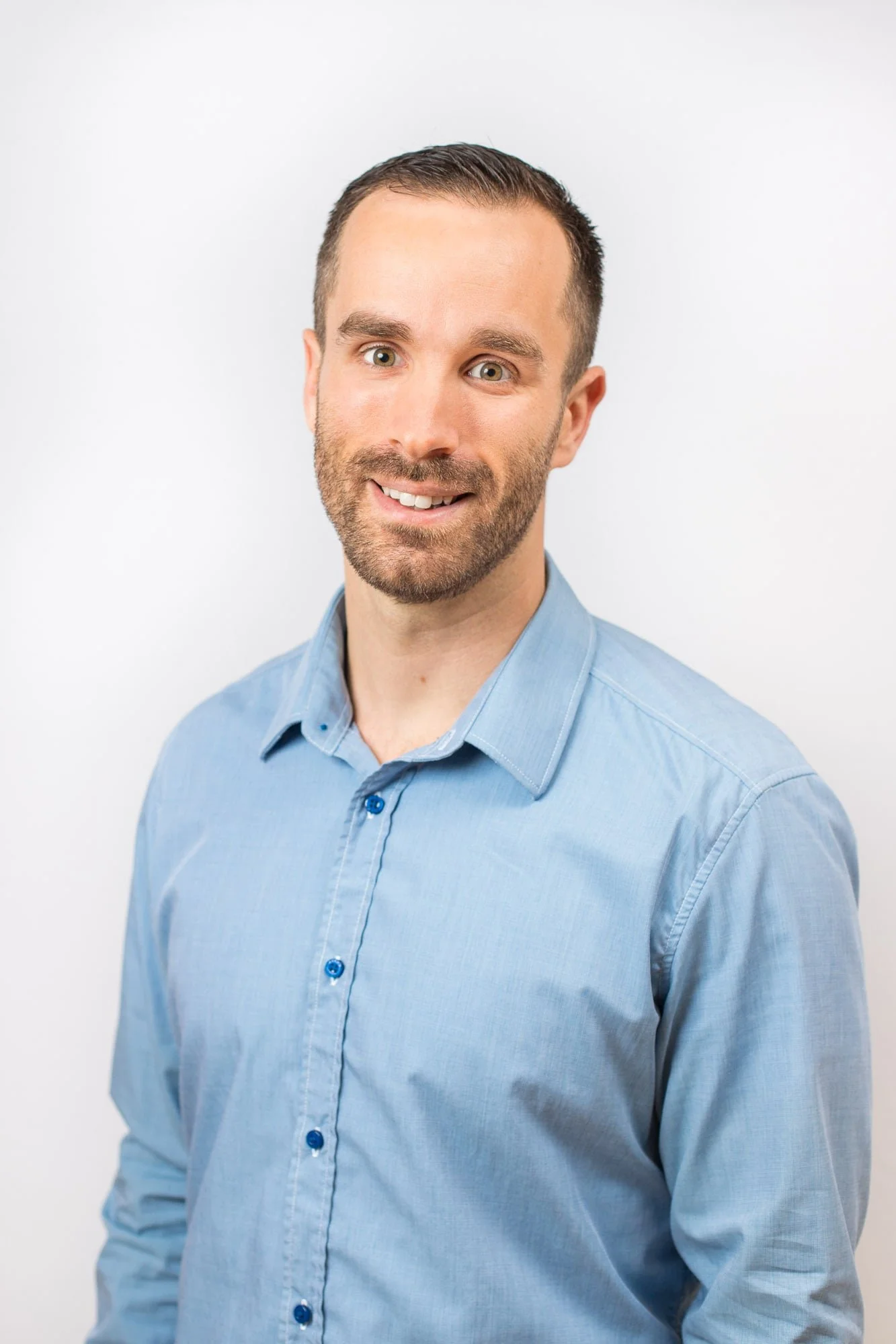 A man with short brown hair, beard, smiling, wearing a light blue button-up shirt, standing against a plain white background.
