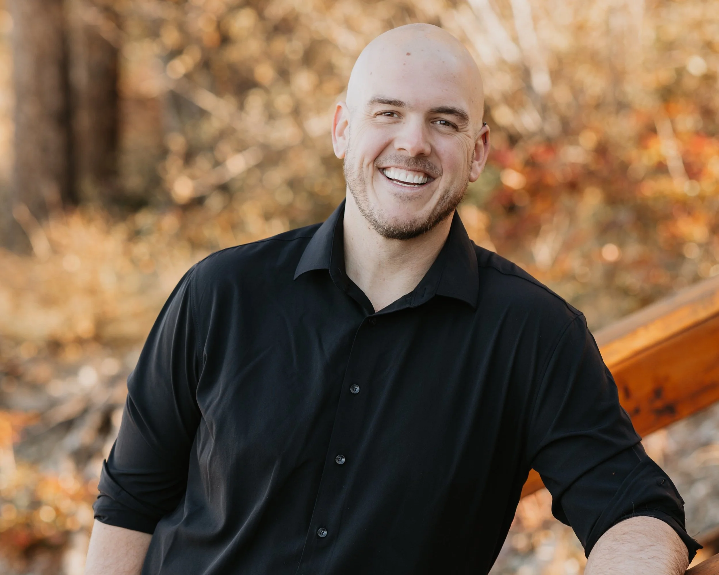 A smiling bald man in a black button-up shirt standing outdoors with autumn foliage in the background.
