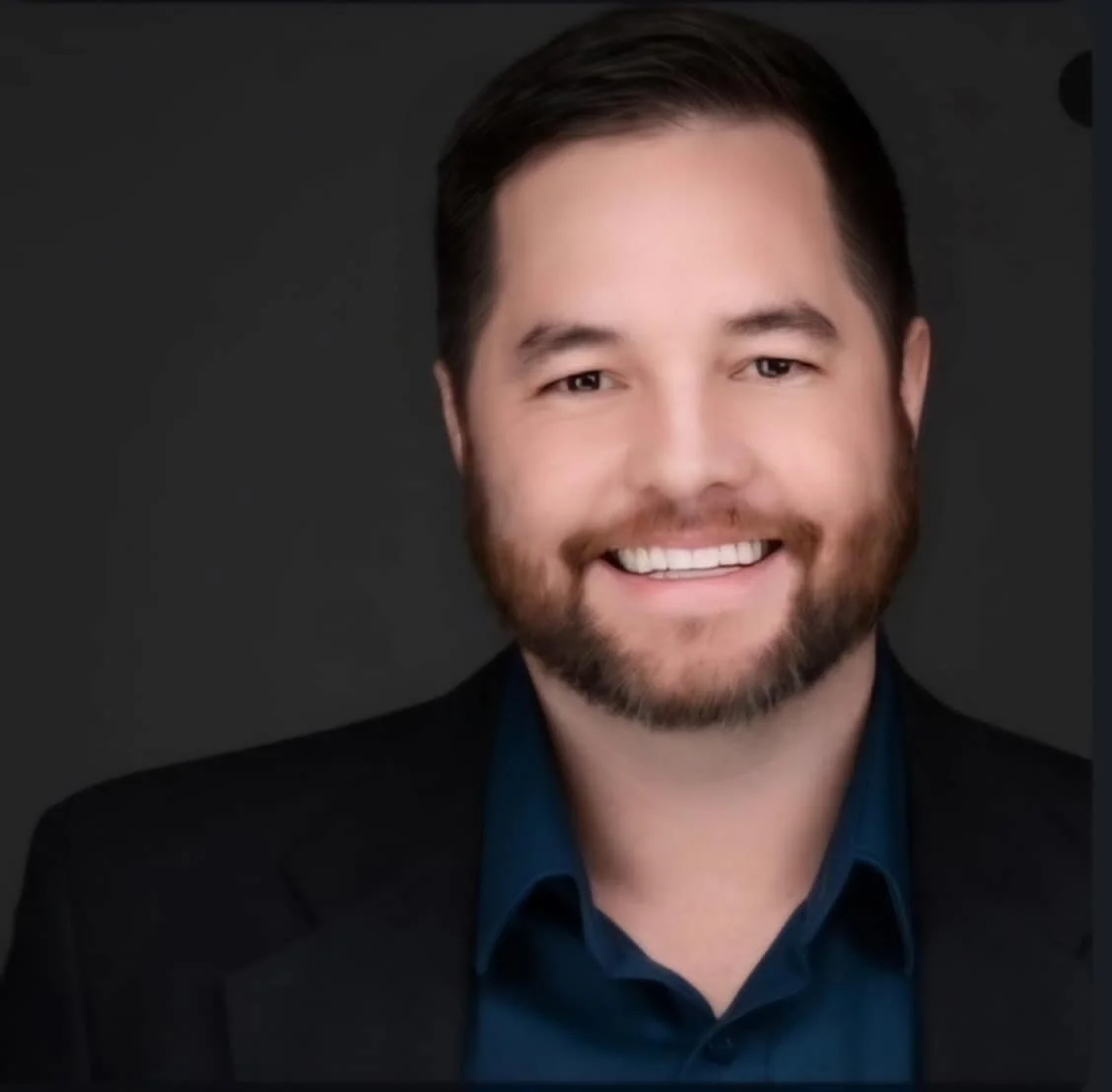 Headshot of a smiling man with dark hair, beard, wearing a navy blue shirt and black blazer against a dark background.